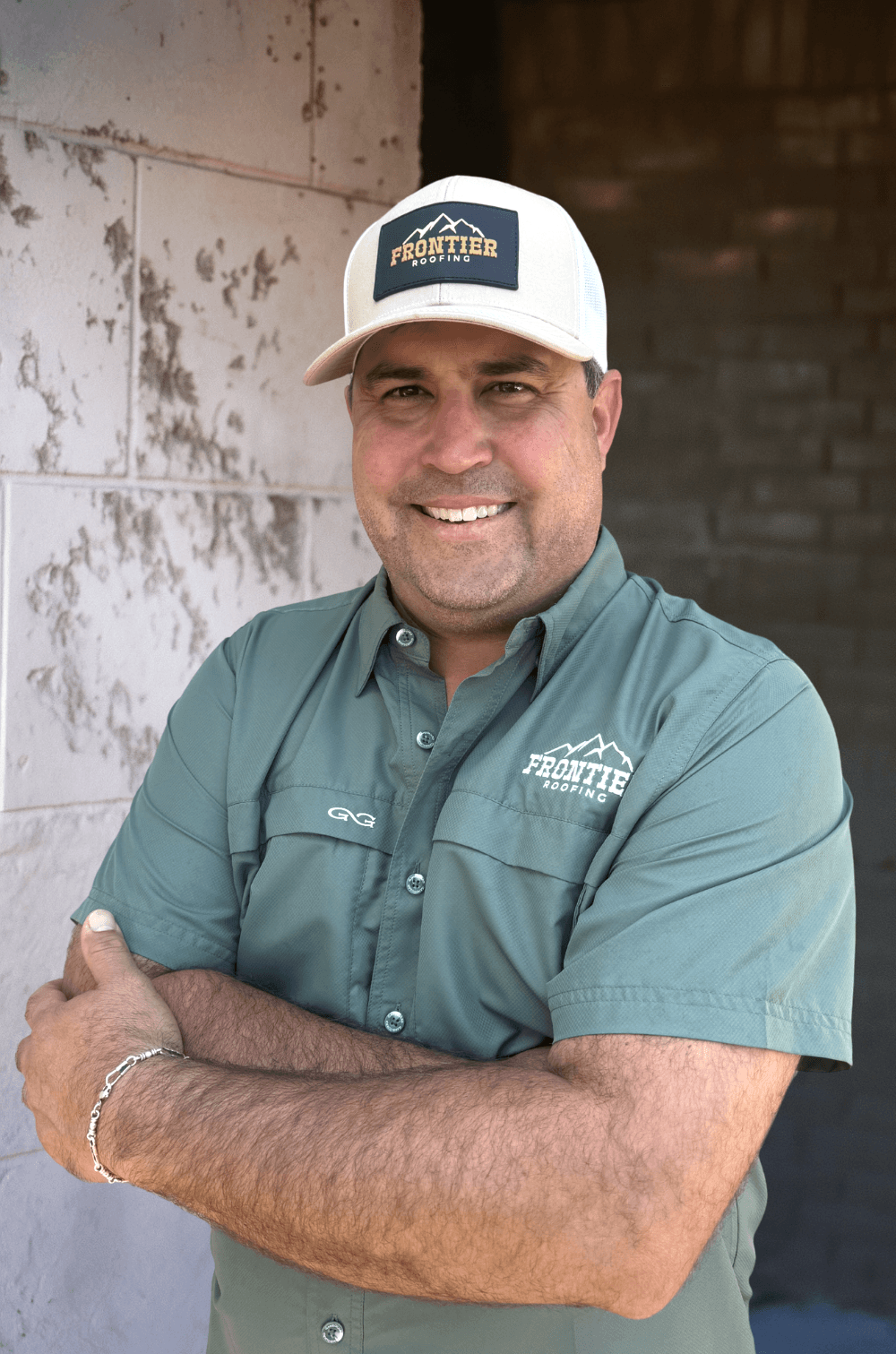 A man smiling with arms crossed, wearing a green collared shirt and white baseball cap with 'Frontier Roofing' logo, standing against a background of a white and brown wall.