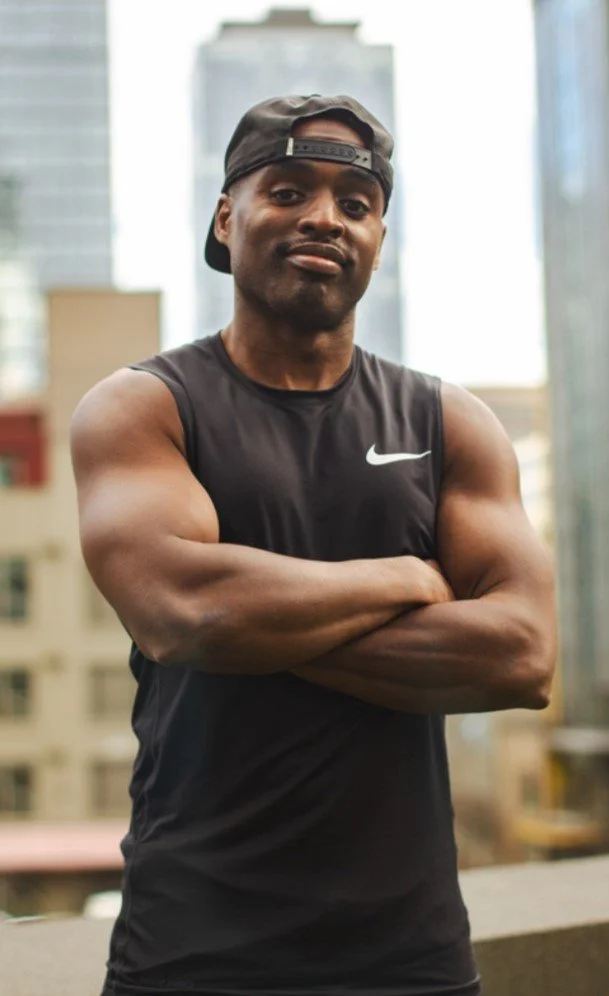 A young man in athletic wear with crossed arms standing outdoors in an urban setting with tall buildings in the background.