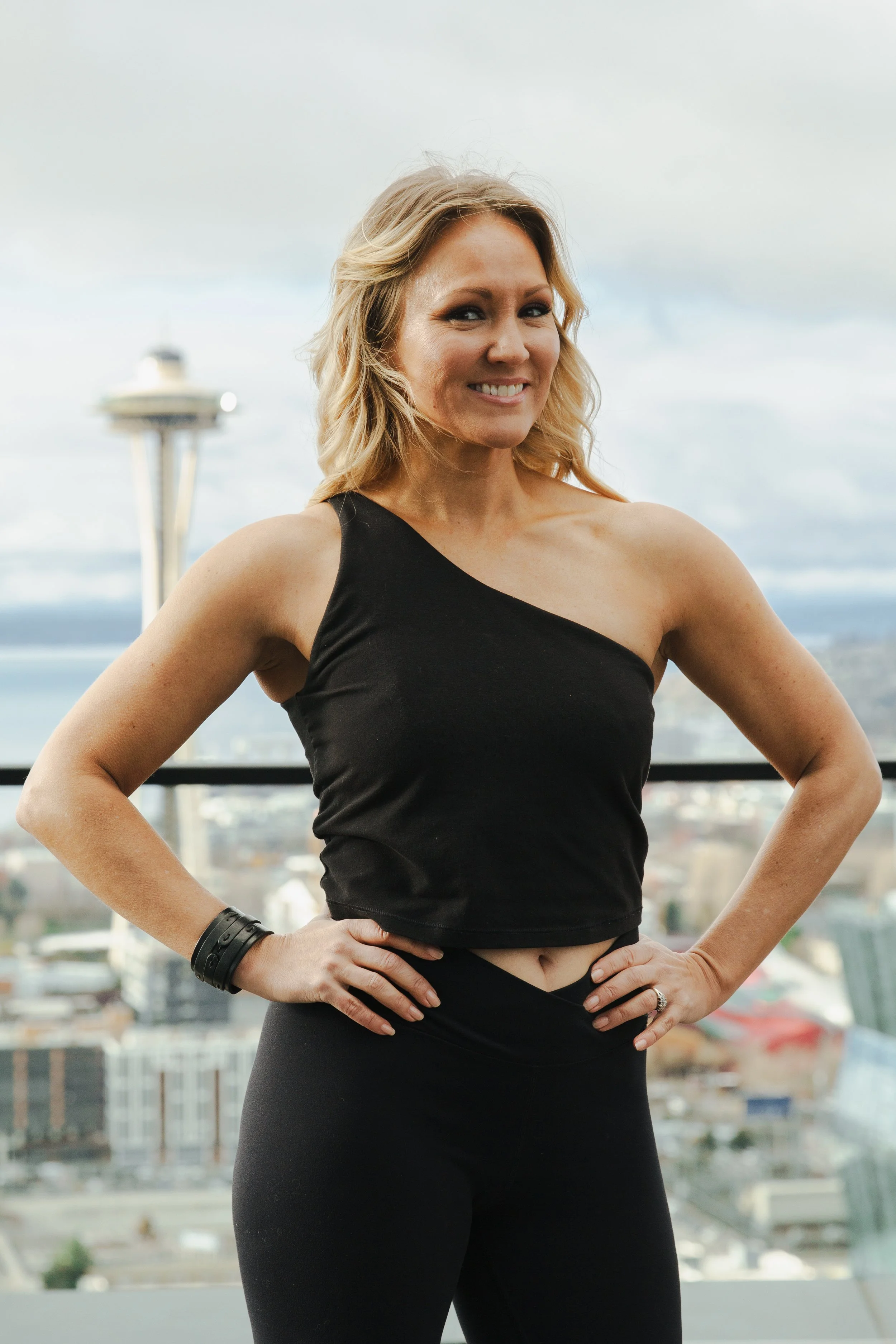 A woman with blonde hair smiling, standing with hands on hips, wearing a black sleeveless top and black leggings, with the Space Needle and cityscape in the background
