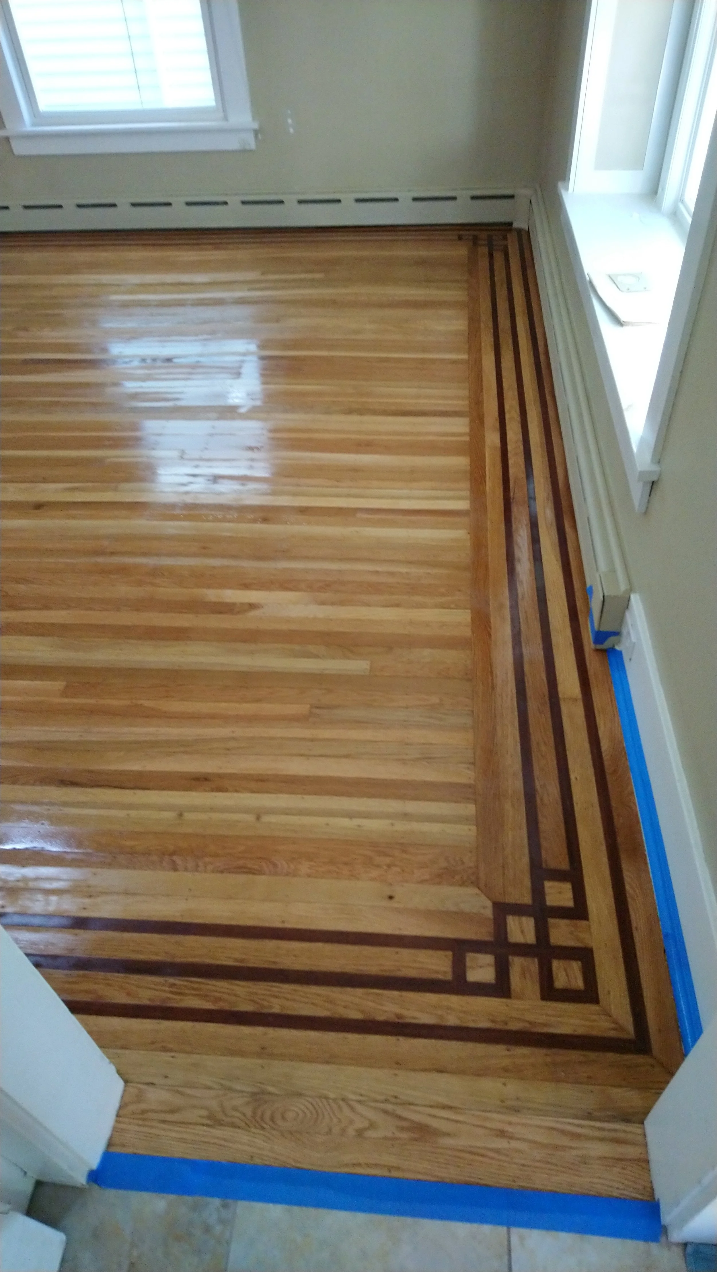 Close-up of a hardwood floor with a decorative border pattern, near a white door frame and a window, with blue painter's tape along the edge.