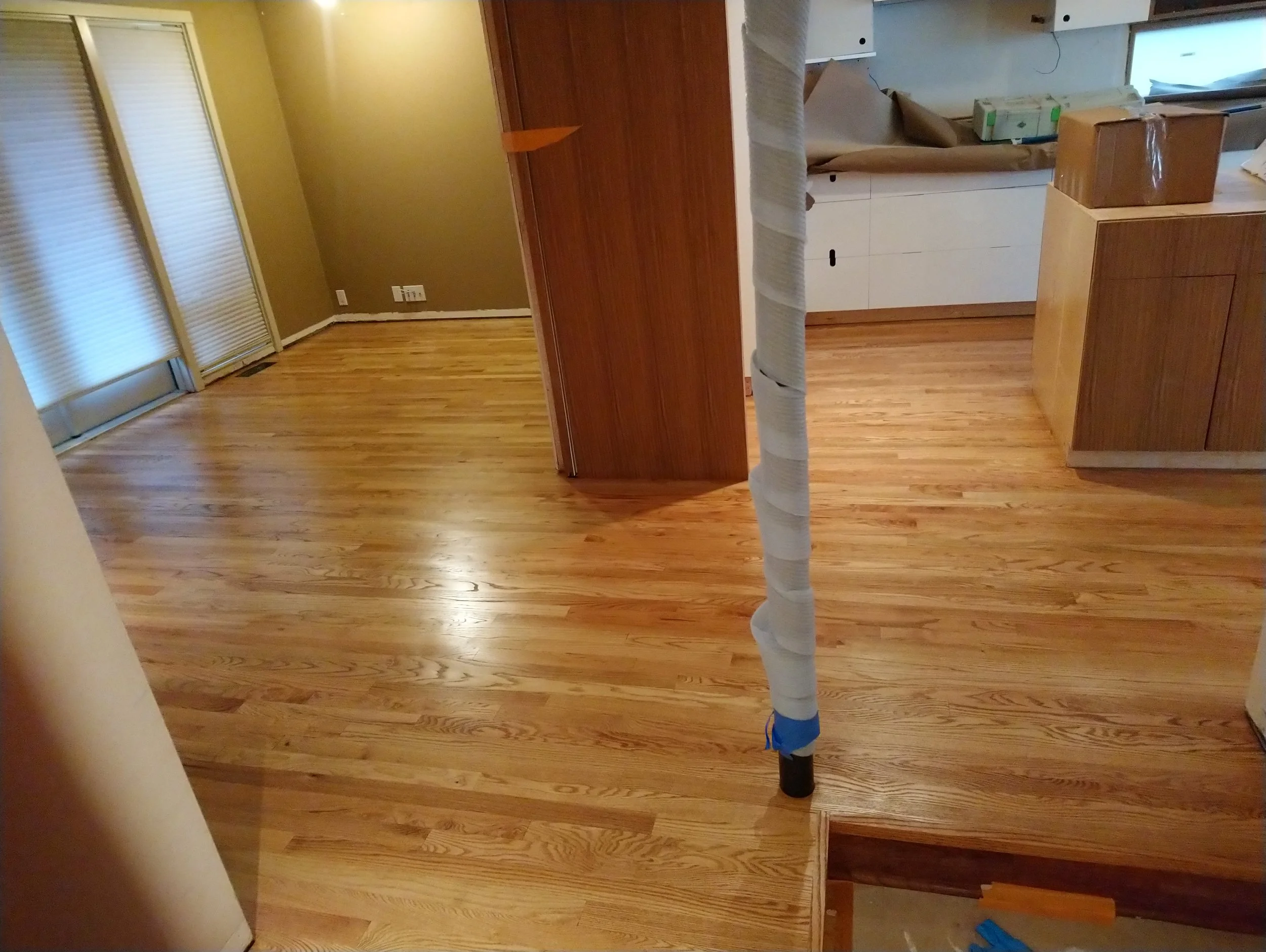 Living room with wooden floors, yellow walls, and sliding glass doors with white blinds. There are some boxes and a piece of furniture covered in protective paper next to a large duct pipe.