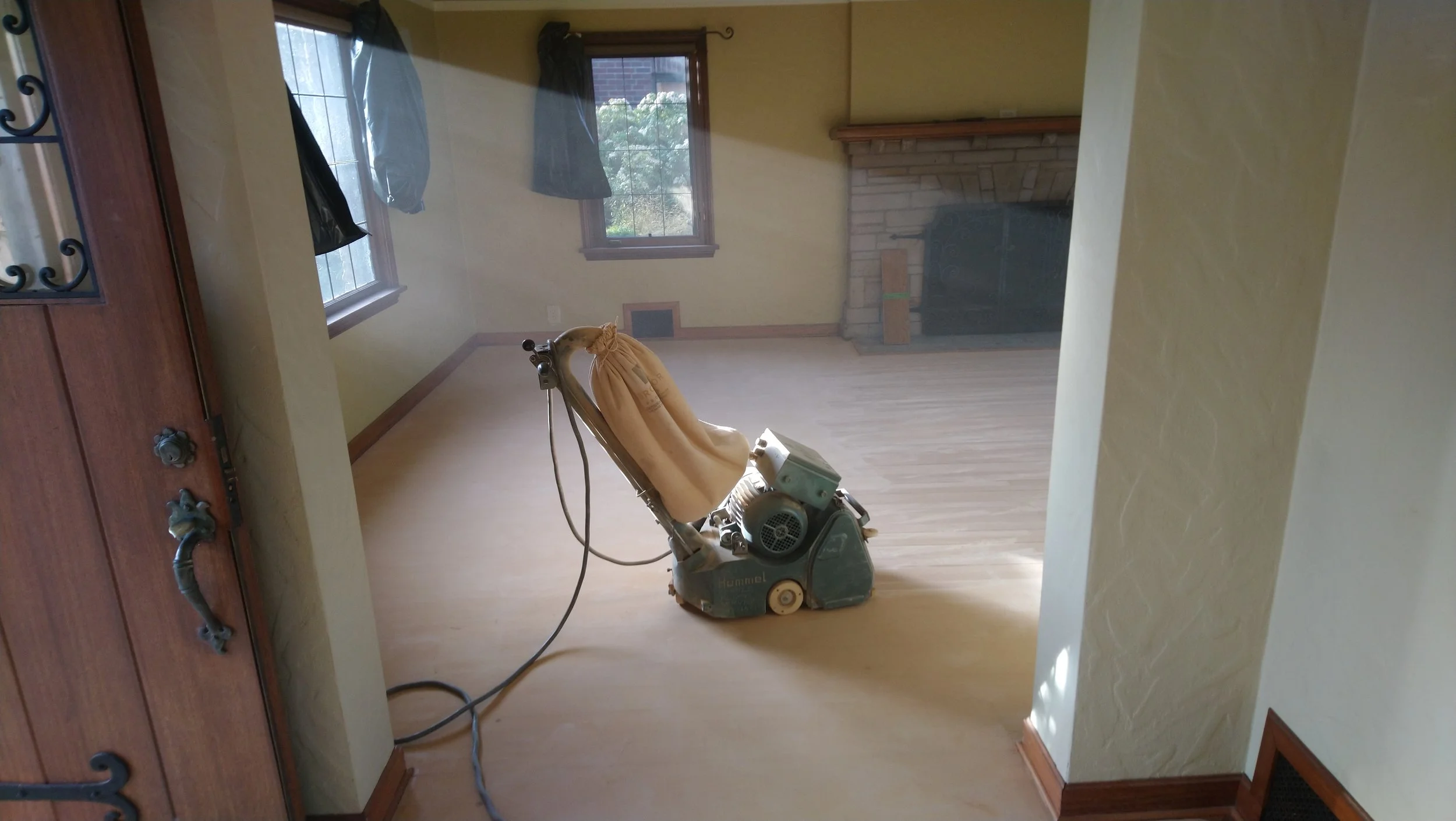 A room undergoing renovation with a floor polishing machine, double-hung windows, fireplace, and hardwood floors.