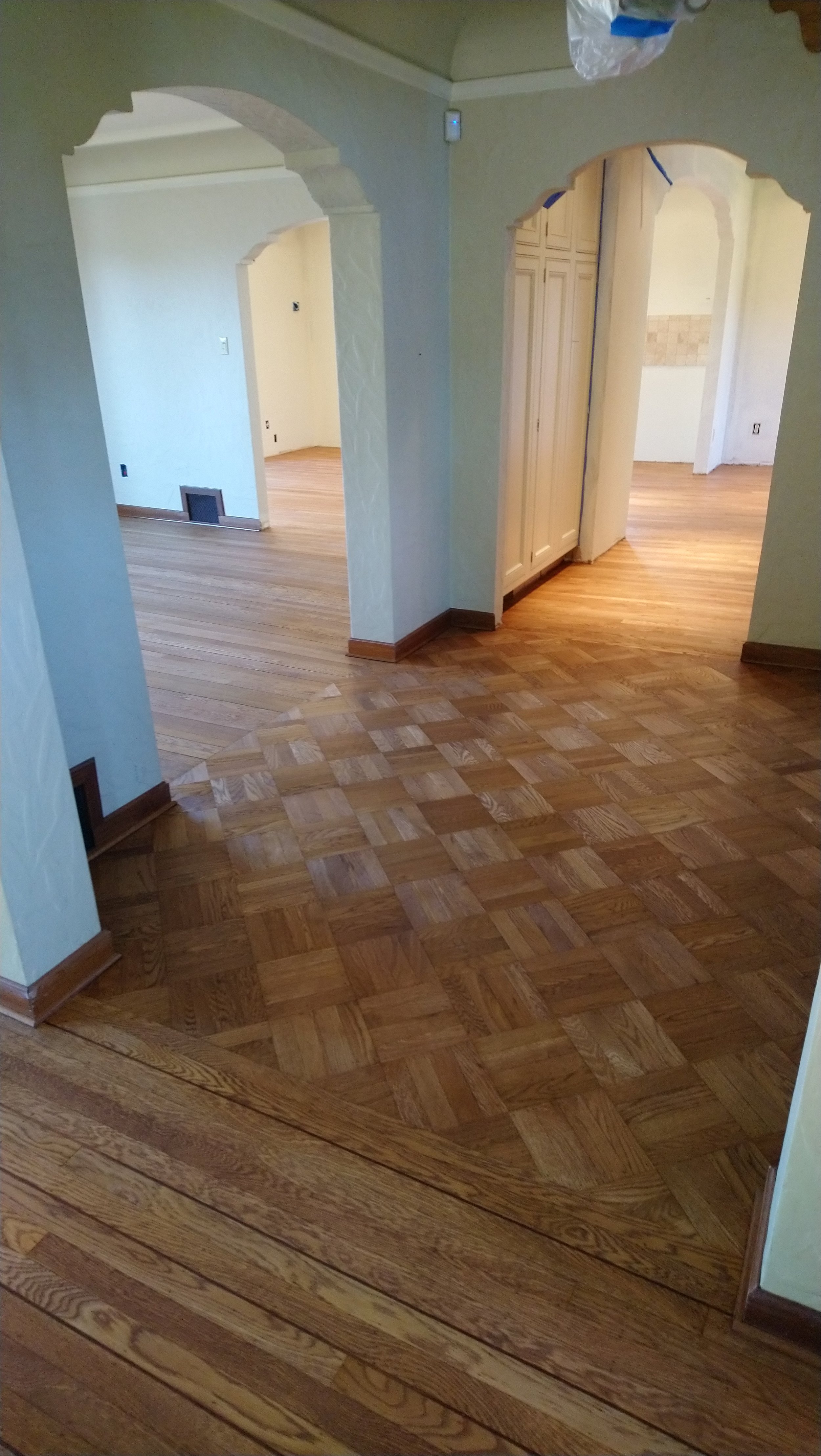 Interior view of a house with multiple rooms, showcasing a hardwood parquet floor pattern with a glossy finish, light-colored walls, arched doorways, and a small area with wood cabinetry. There is also a wall vent and a hanging plastic bag visible.