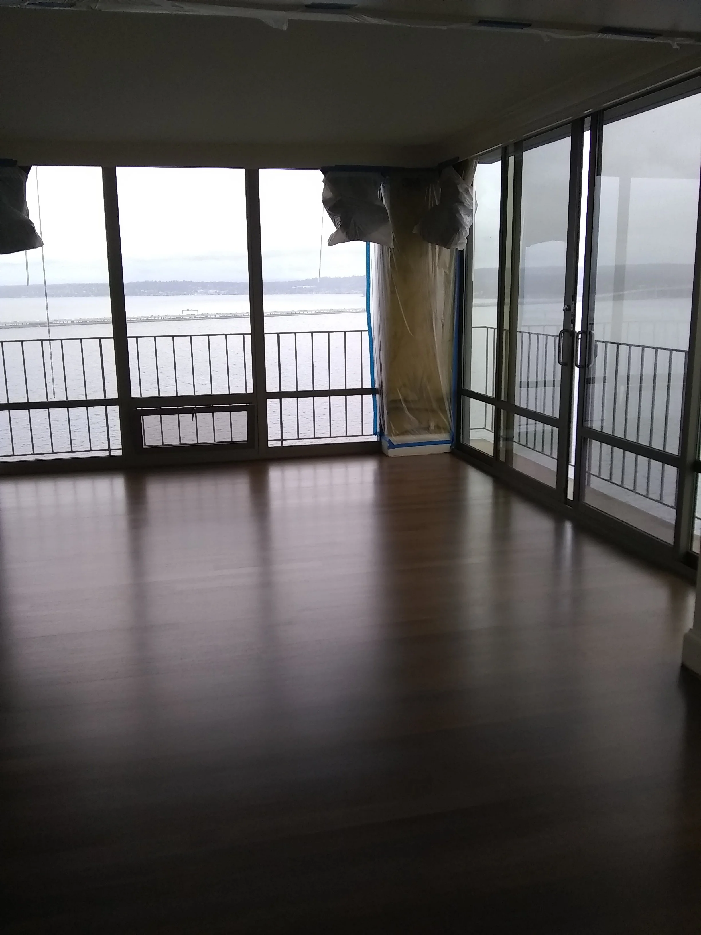 Interior view of a room with wooden floors, large windows, and a balcony overlooking water. There are two covered objects near the windows, and some of the window panes are open.