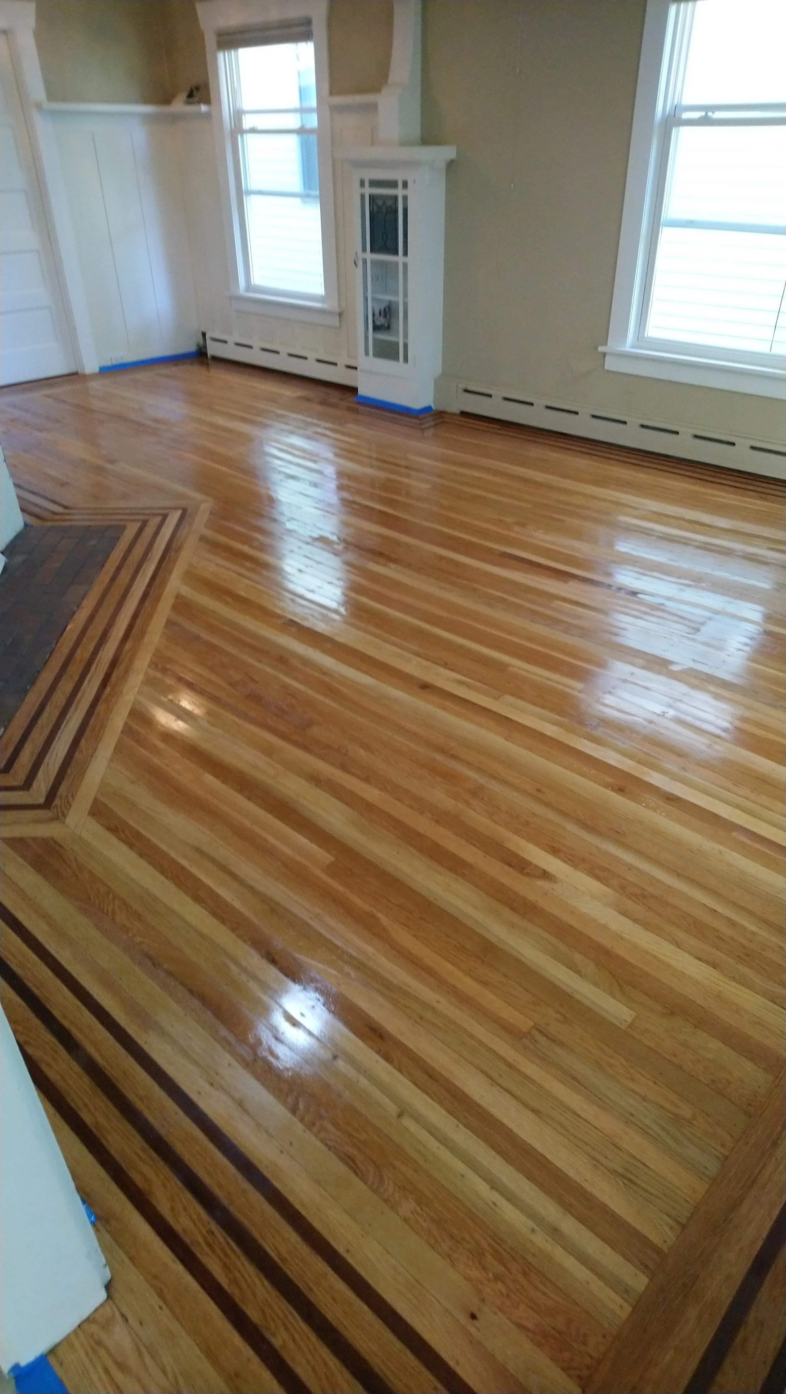 Refinished hardwood floor in a living room with two windows, a small built-in display cabinet, and baseboard heating.