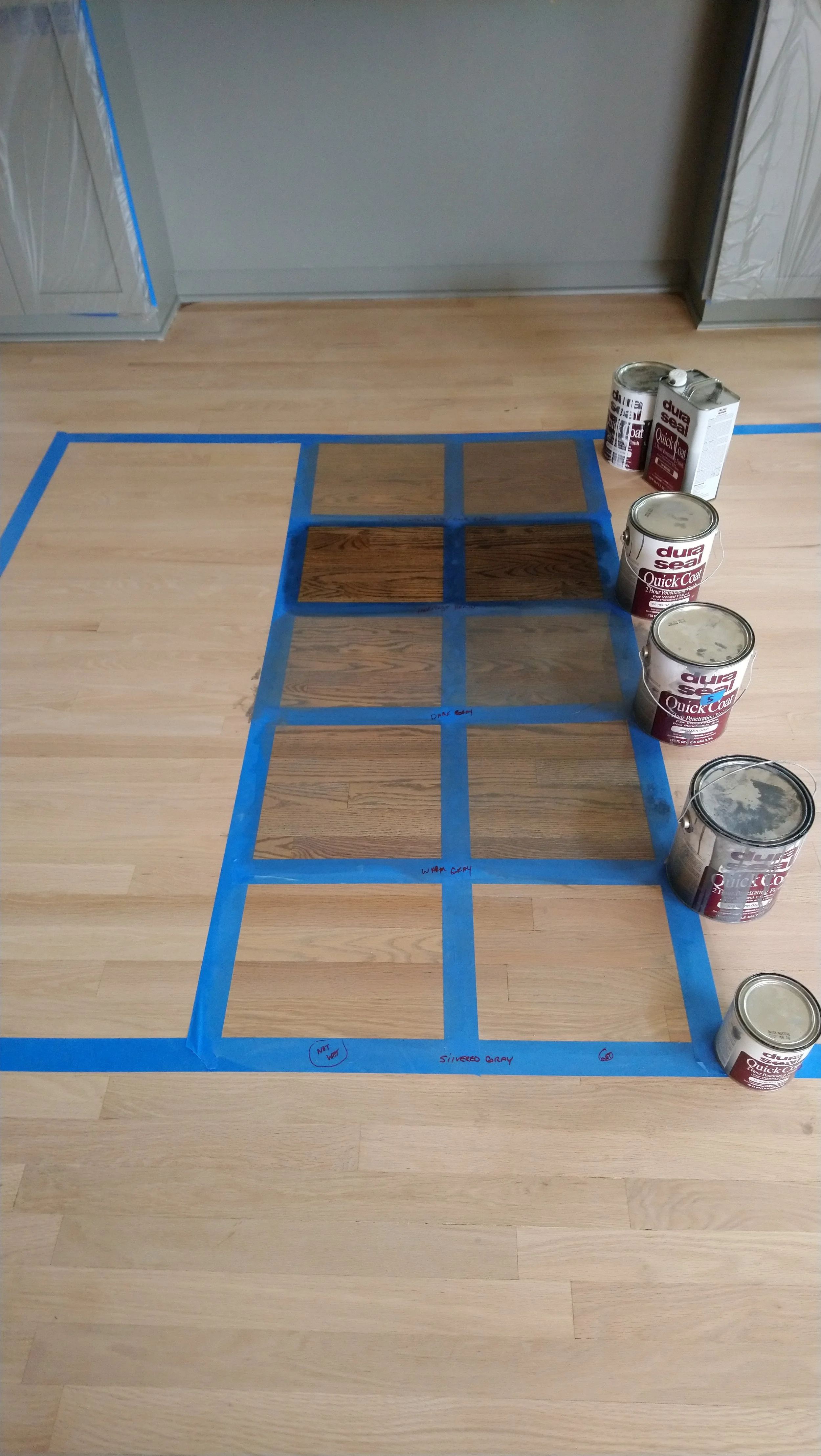 A hardwood floor with blue painter's tape creating a grid pattern. Several cans of wood stain are placed on the floor to the right of the taped area, along with a metal container of wood stain. The tape appears to designate sections for staining or p