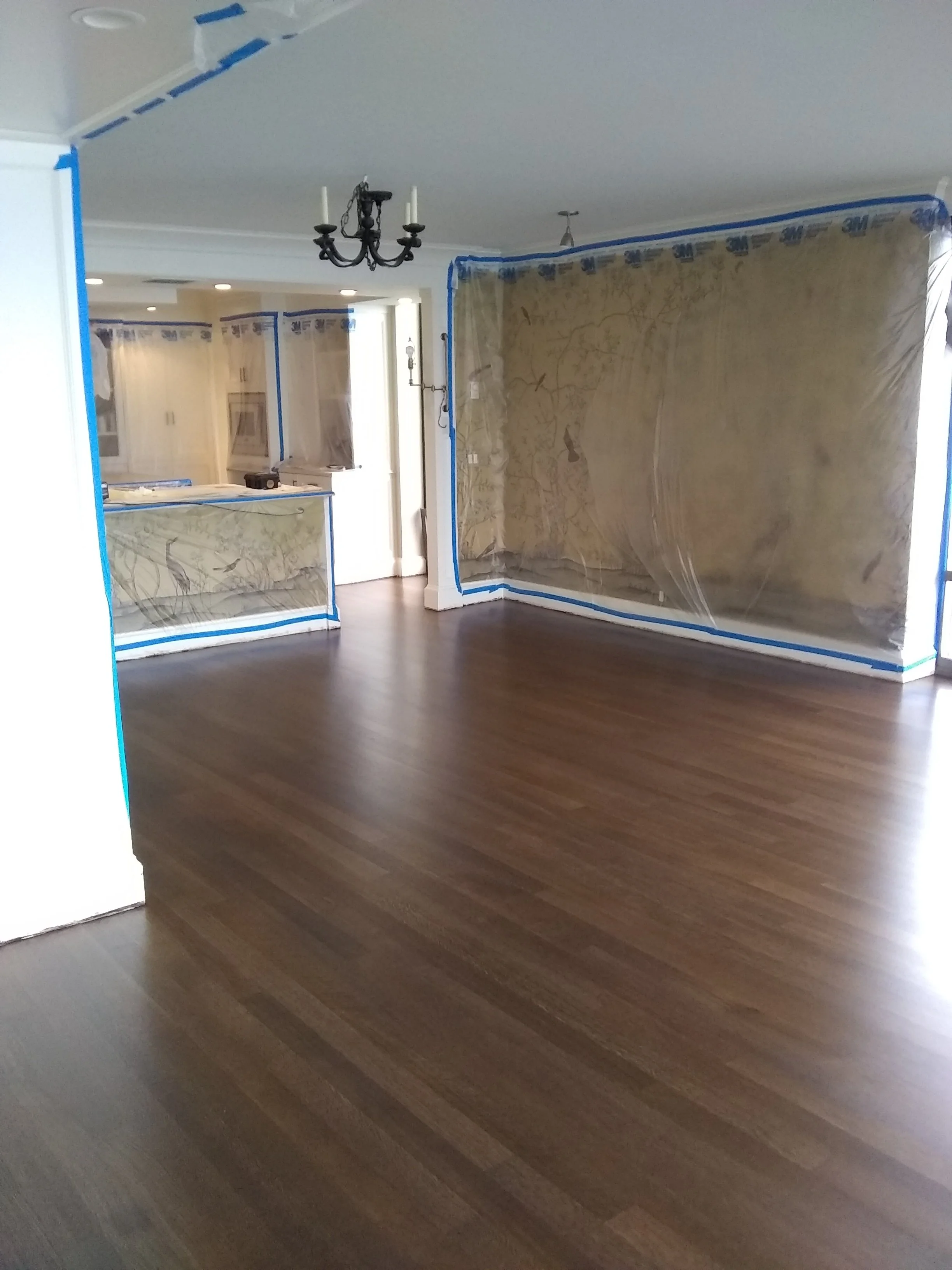 Interior of a house under renovation, with walls partially covered with plastic sheeting and blue painter's tape, and a chandelier hanging from the ceiling.