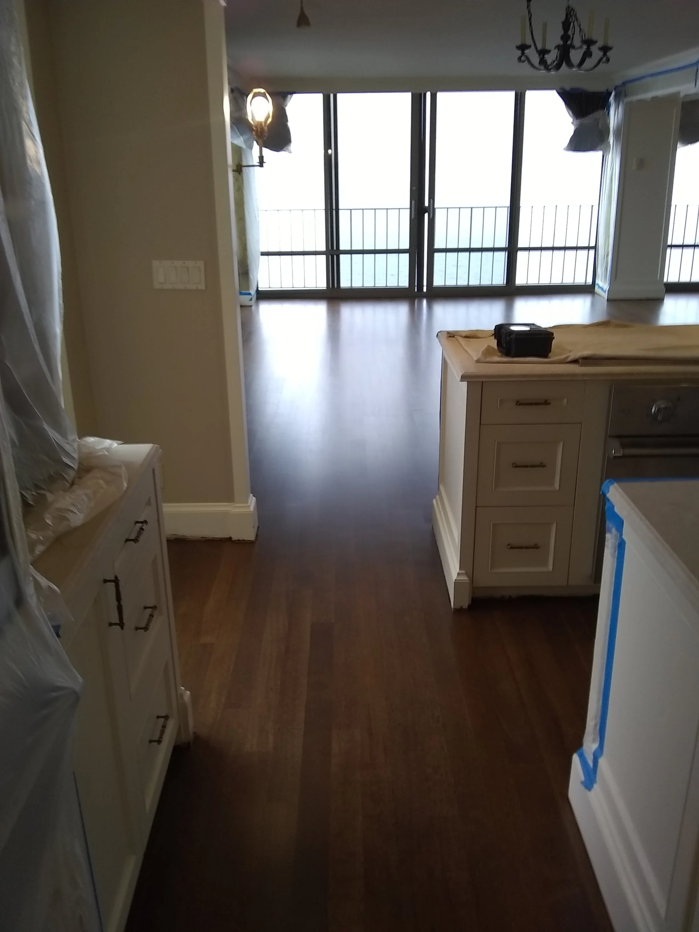 An interior view of a room under construction or renovation, showing dark hardwood floors, white cabinets, a chandelier, and a large window or sliding door overlooking a body of water.