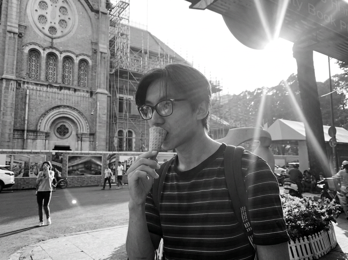 A young man wearing glasses and a striped shirt is eating an ice cream cone outdoors in front of a historic building covered with scaffolding. The photo is in black and white, and the sun is shining brightly.