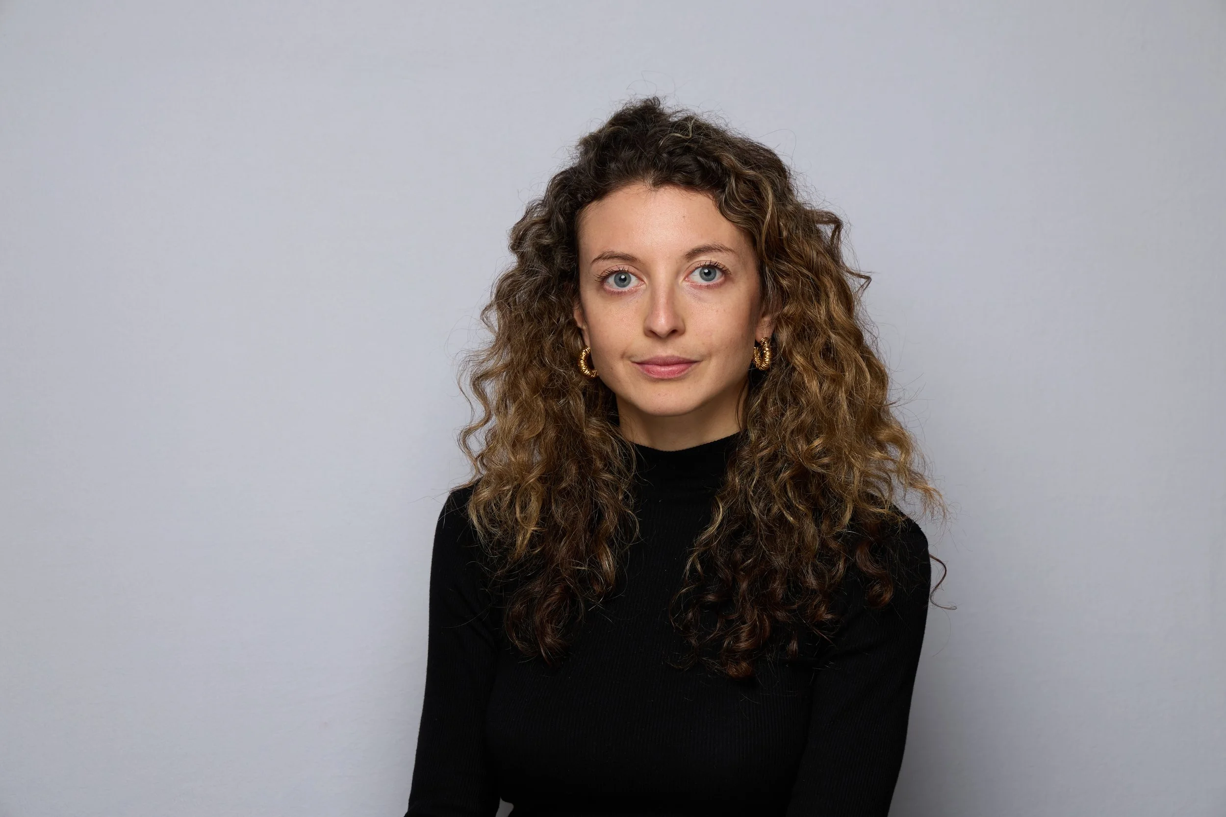A woman with curly brown hair, blue eyes, wearing a black turtleneck and gold hoop earrings, looking at the camera against a plain gray background.