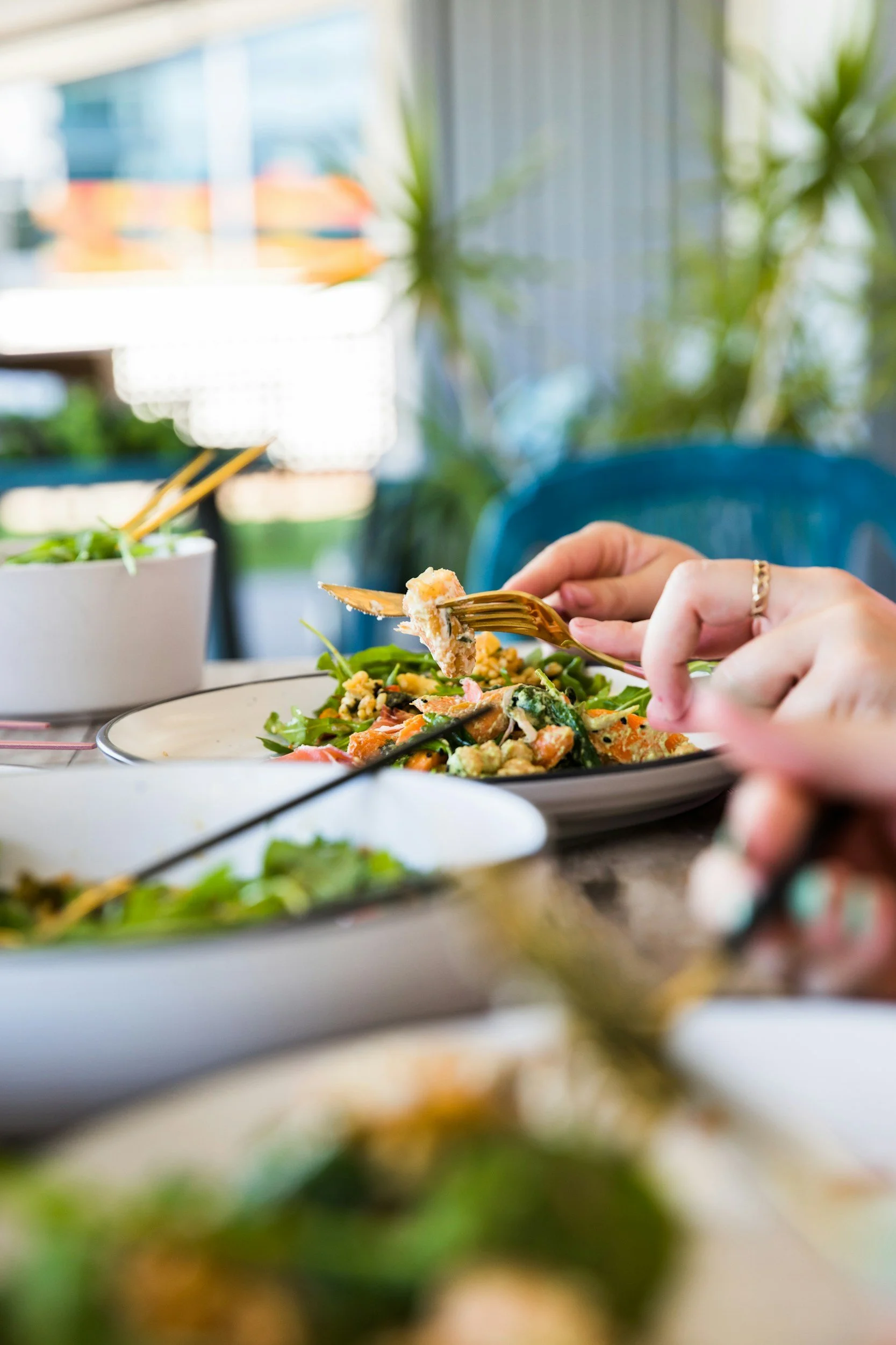 Person eating a salad with a fork at a dining table, with a plant and window in the background.