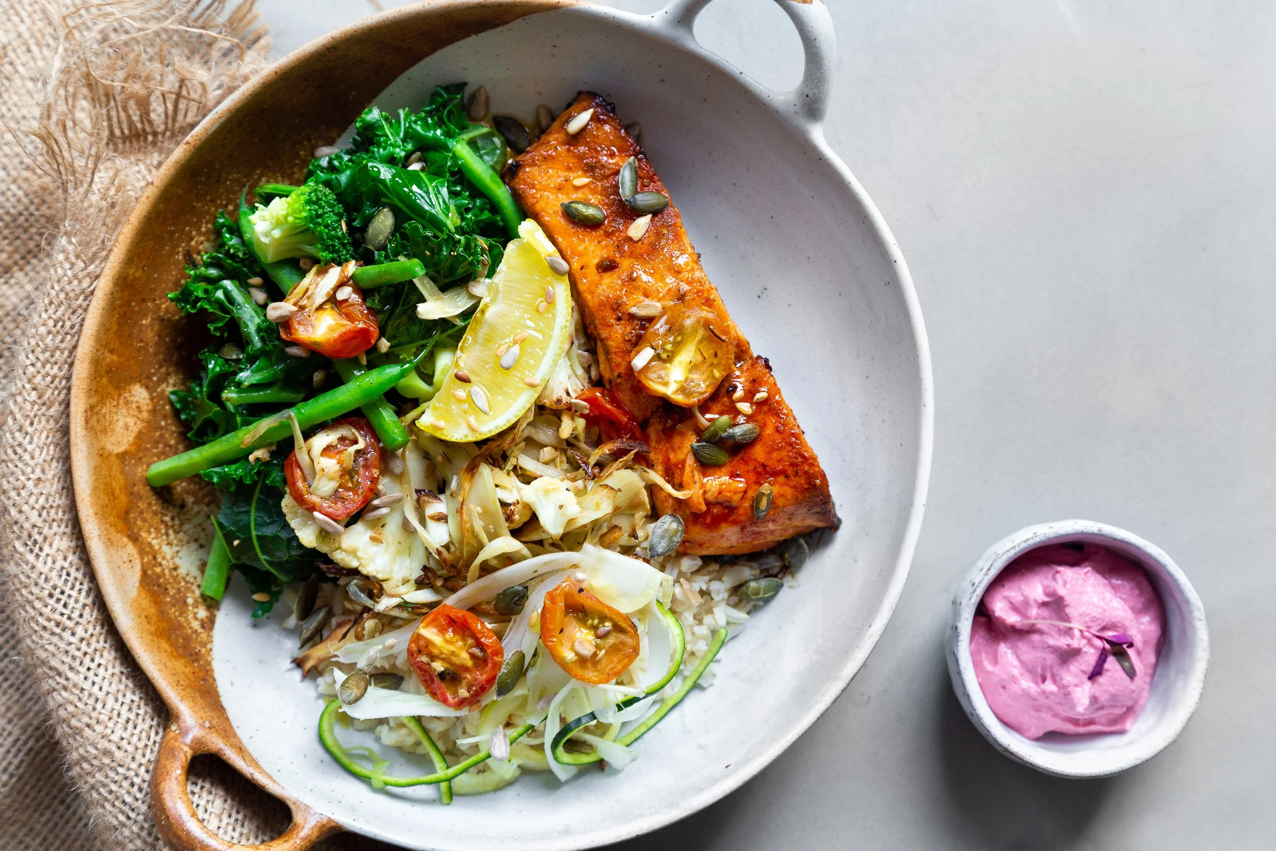 A plate with a salmon fillet, lemon wedge, and vegetables including broccoli, cherry tomatoes, zucchini, cauliflower, and leafy greens, with a small bowl of pink sauce on the side.