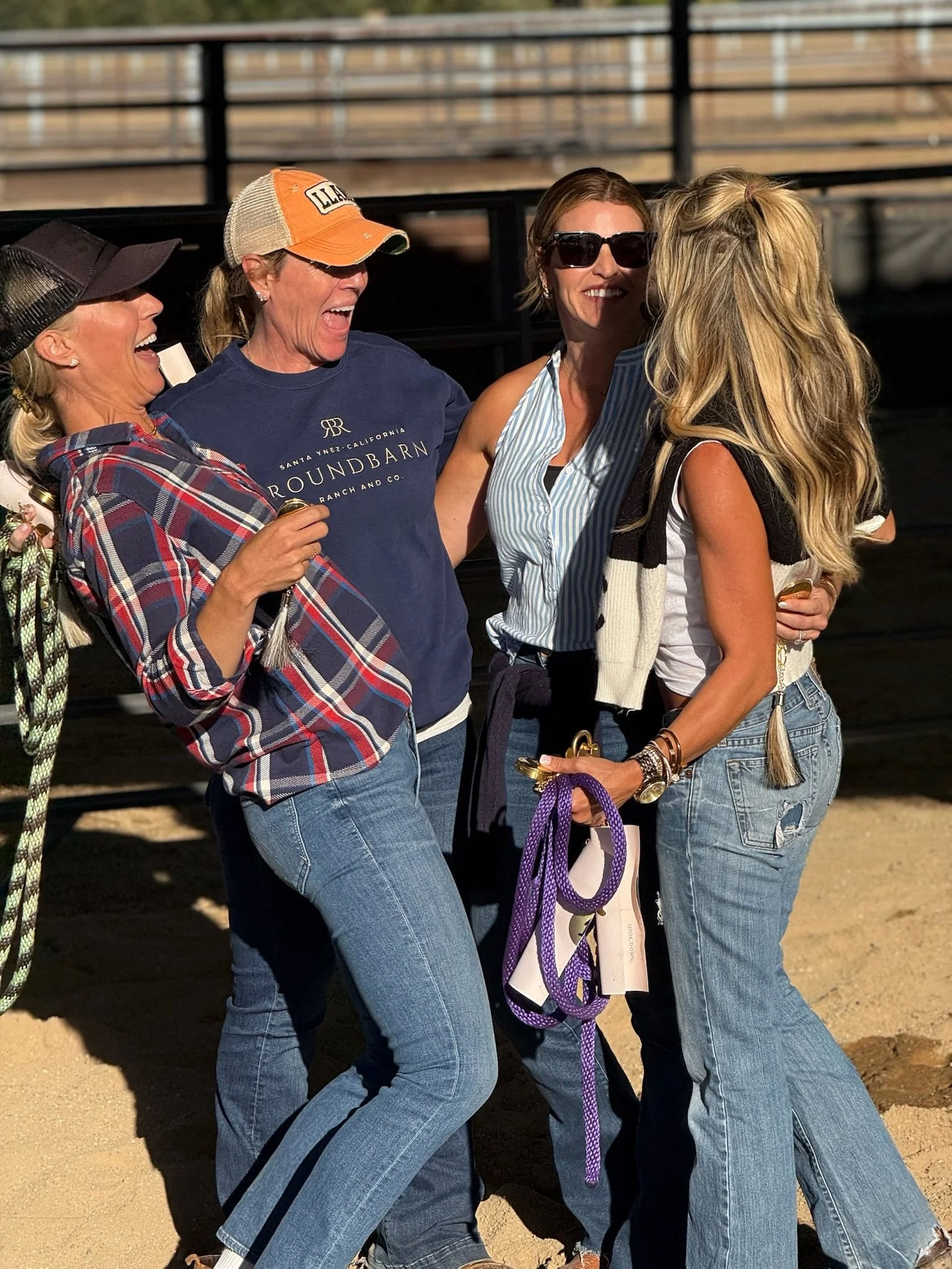 Four women wearing casual clothing and sunglasses are laughing and hugging on a sunny day near a wooden fence.