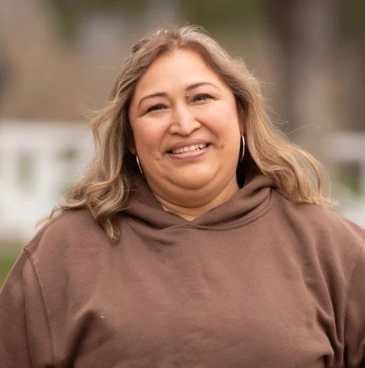 A woman with shoulder-length blonde hair, smiling, wearing a brown hoodie, outdoors with blurred greenery in the background.