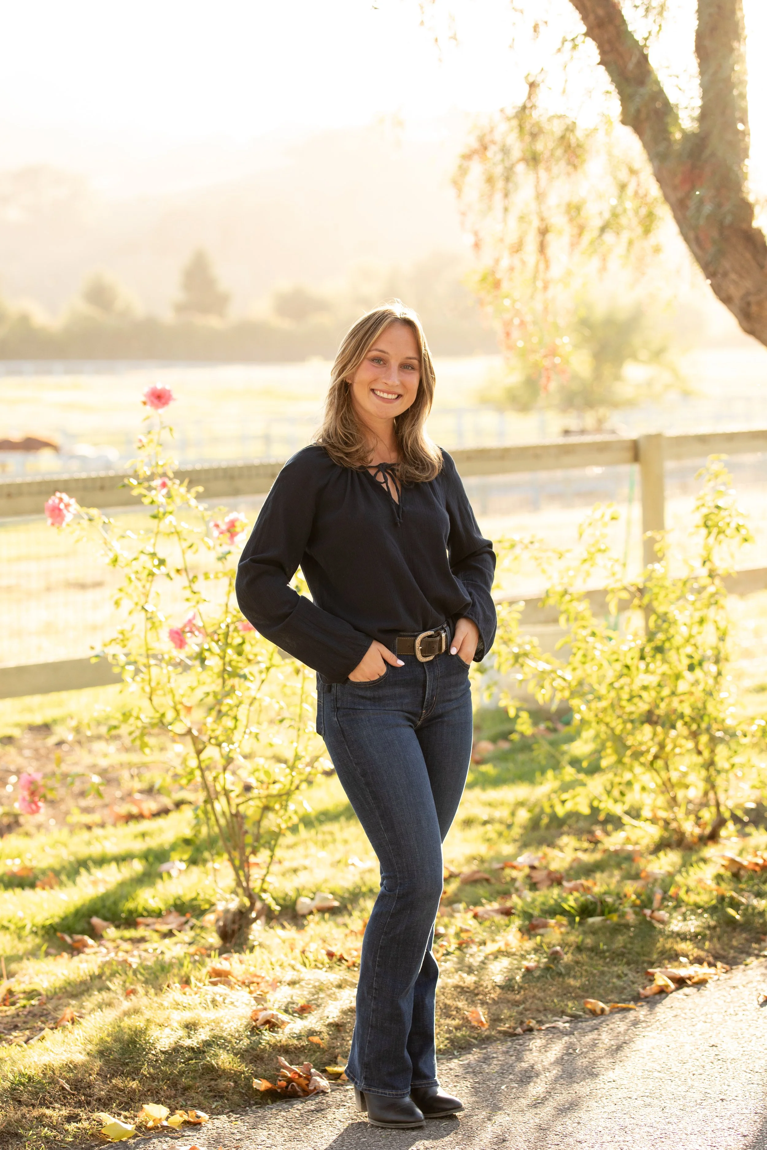 A woman stands outside near a large tree, smiling, with sunlight casting a warm glow, surrounded by flowers and greenery.