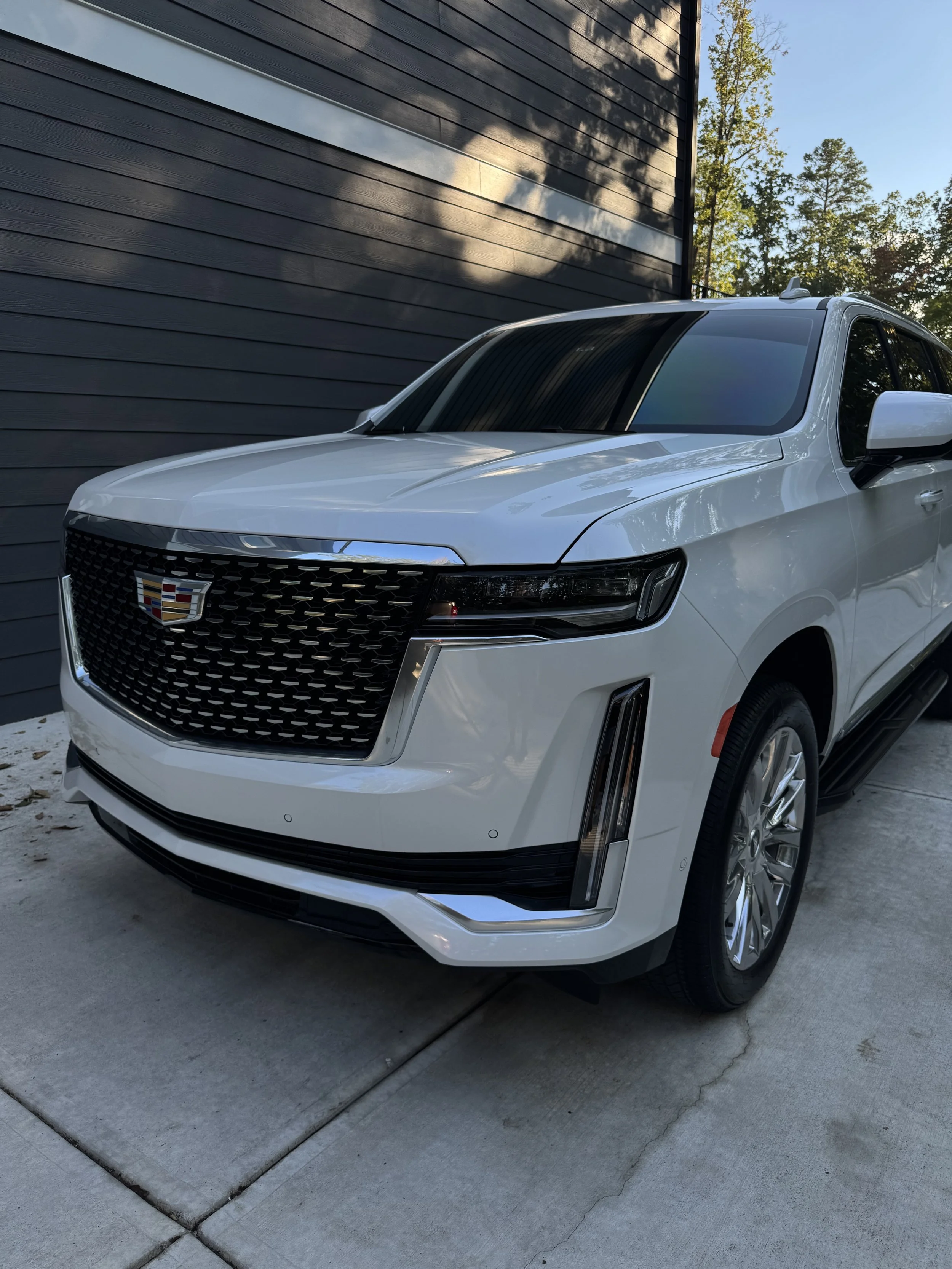 A white Cadillac SUV parked in front of a black house with horizontal siding, trees in the background, during daytime.