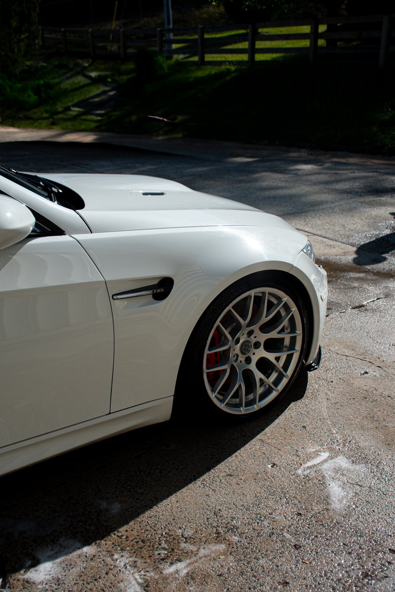 Close-up of a white sports car, focusing on the front wheel and part of the hood, parked on a weathered concrete surface with sunlight and shadows.