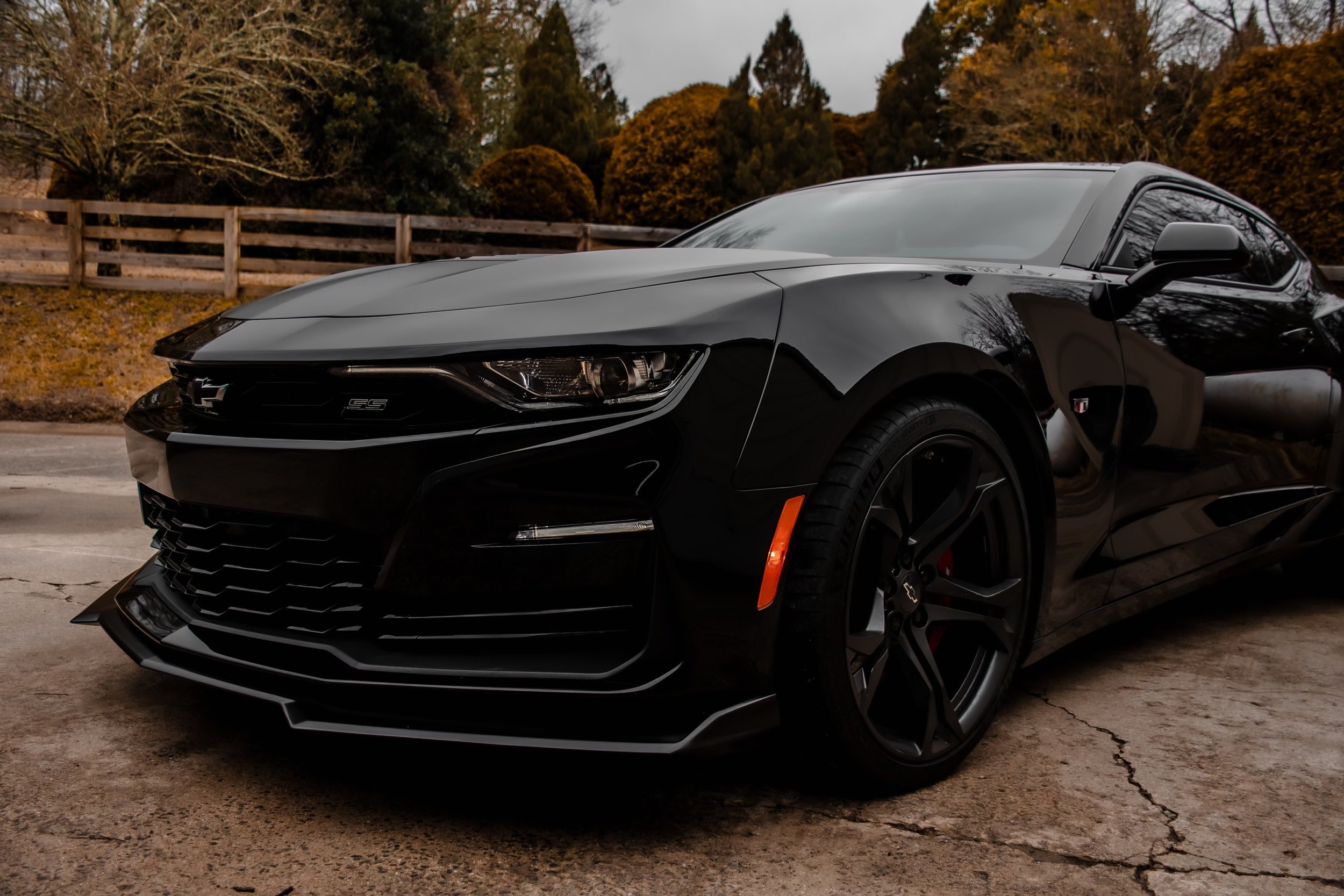 A black Chevrolet Camaro SS sports car parked on a concrete surface with a wooded area and wooden fence in the background.