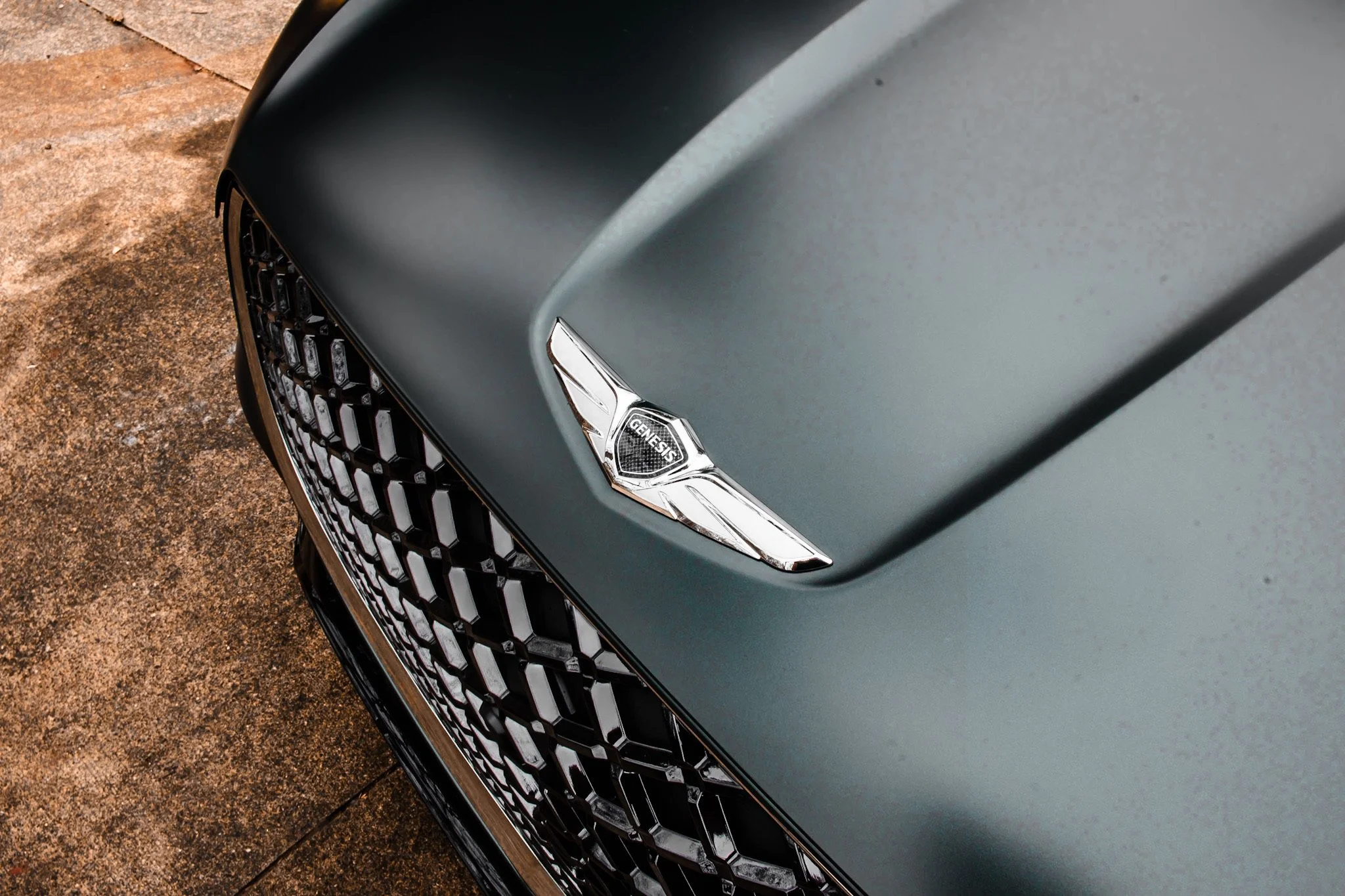 Close-up of the front end of a dark gray Genesis car, focusing on the grille, headlight, and a hood ornament with the Genesis logo.