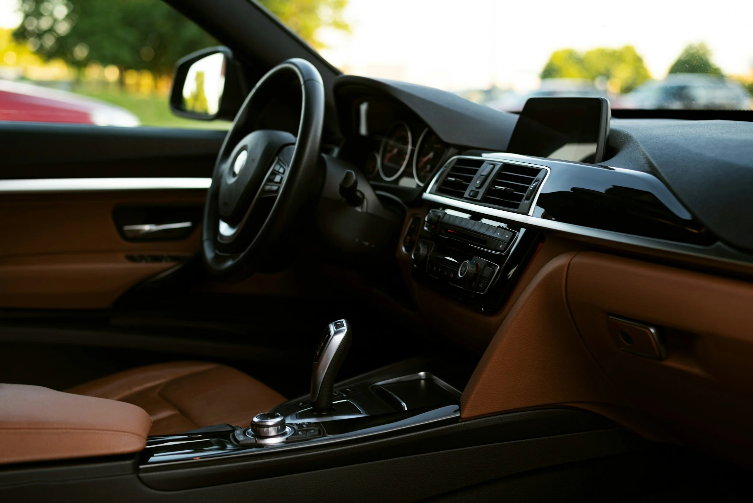 Interior of a car showing the dashboard, steering wheel, gear shift, and center console with climate controls, with a view of trees and other cars outside through the windshield.