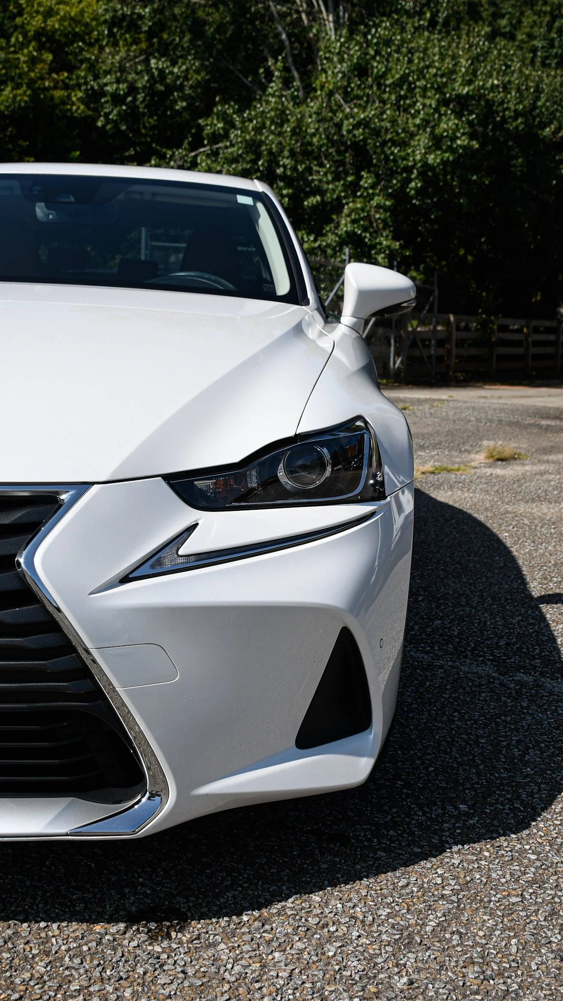 Close-up of the front of a white car parked on a gravel surface with green trees in the background.