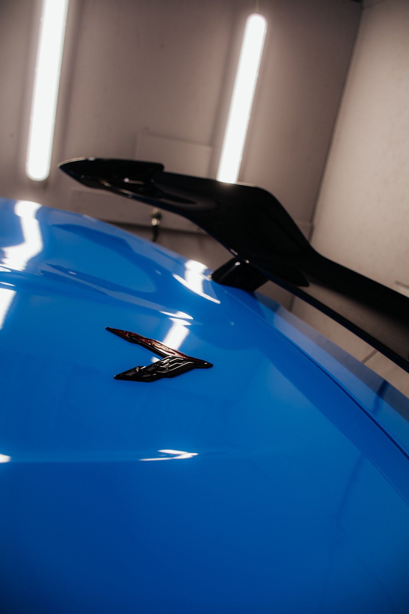 Close-up of a blue sports car with black rear spoiler, showcasing Chevrolet emblem, in a showroom with ceiling lights.