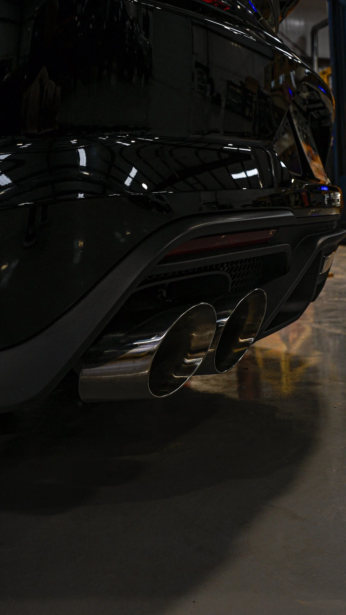 Close-up of the rear of a black sports car showing dual exhaust pipes, part of the rear bumper, and the car's glossy body reflecting the surroundings.