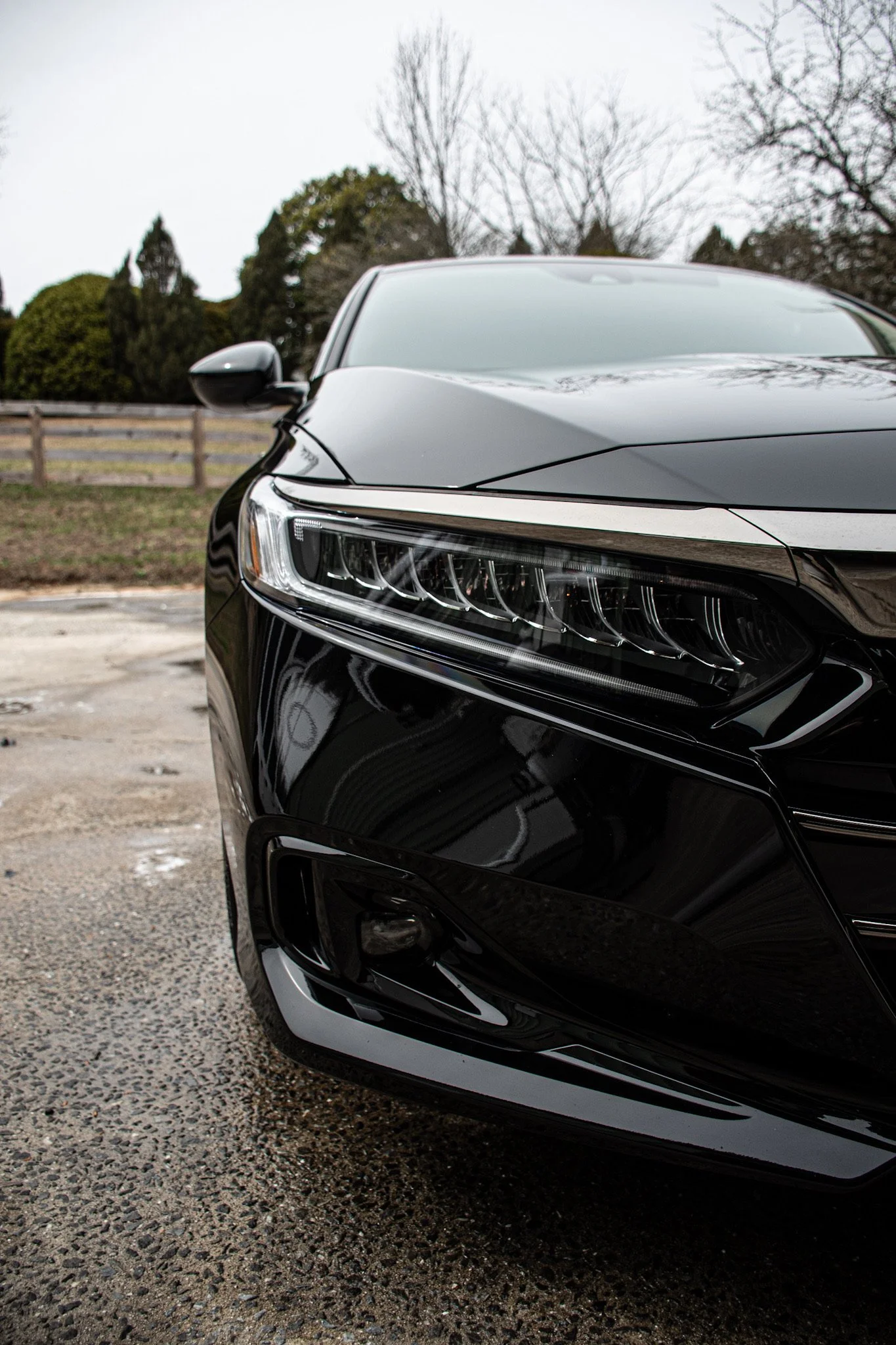 Close-up of the front right side of a black luxury car parked on a gravel surface with trees and a fence in the background.