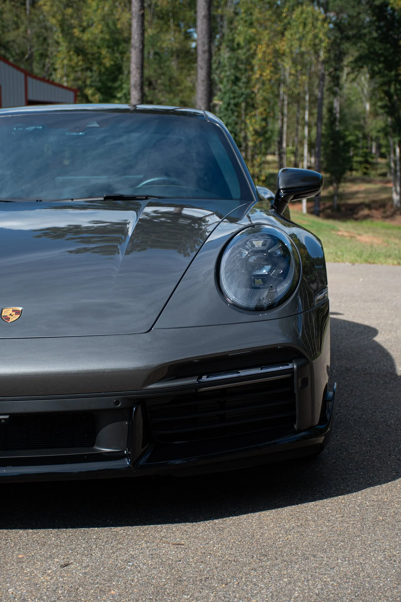 Close-up of the front part of a black Porsche sports car, showing the headlight, logo, and side mirror, with a wooded background.