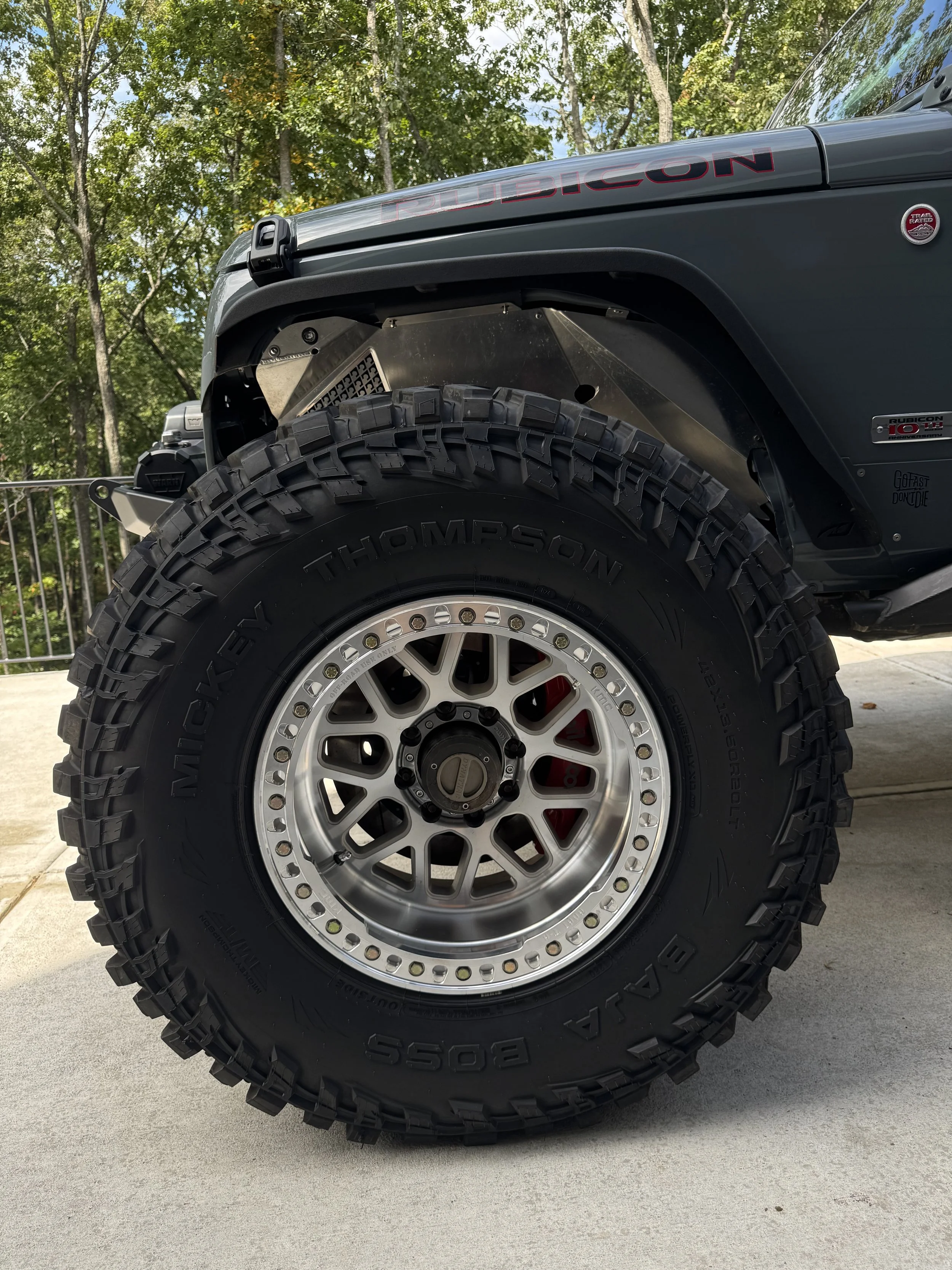 Close-up of a rugged off-road vehicle tire with a silver wheel, parked on a concrete surface with green trees in the background.