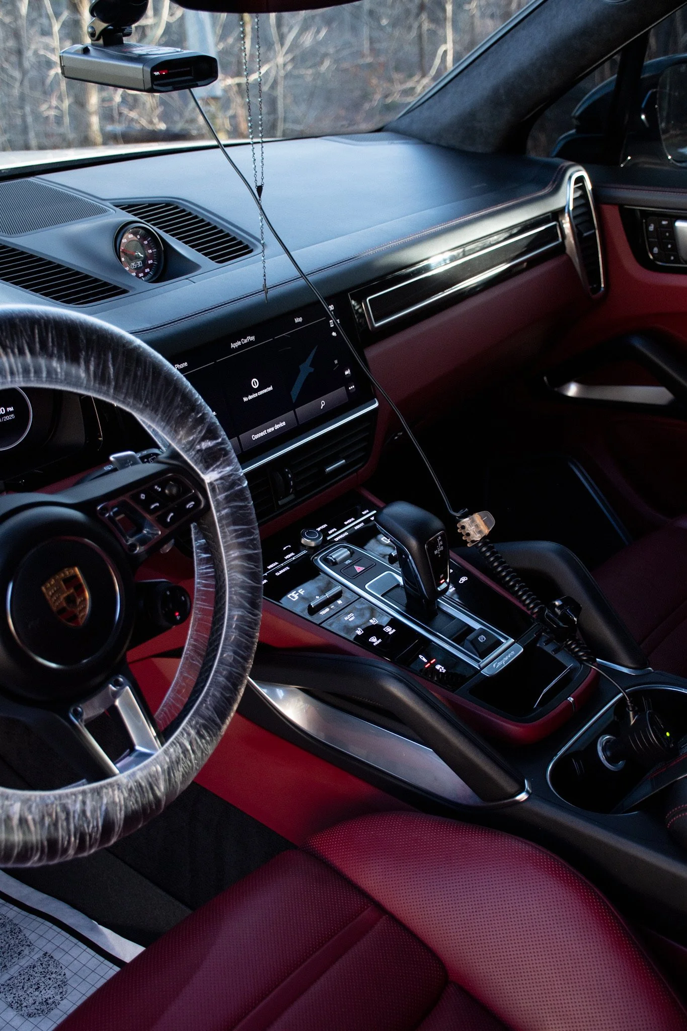 Interior of a luxury car showing a red leather seat, a steering wheel covered with a plastic wrap, a dashboard with a digital display, and a gear shift lever. The background shows the car's windshield and part of the dashboard.