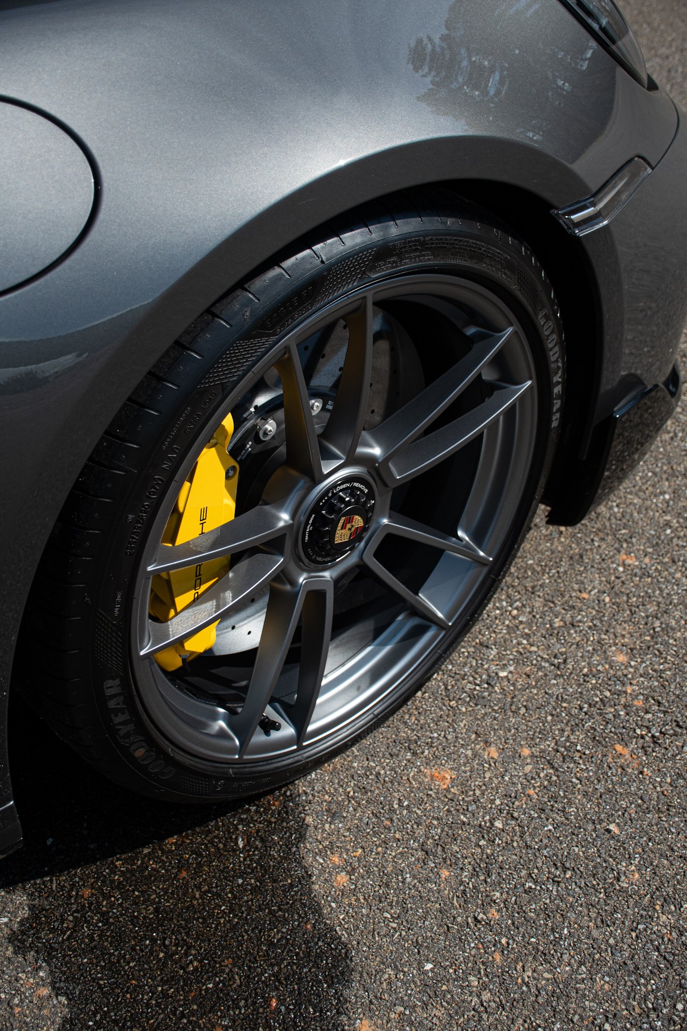Close-up of a gray sports car's front wheel, featuring a black tire, yellow brake caliper, and a multi-spoke alloy rim with a Porsche logo at the center, parked on an asphalt surface.