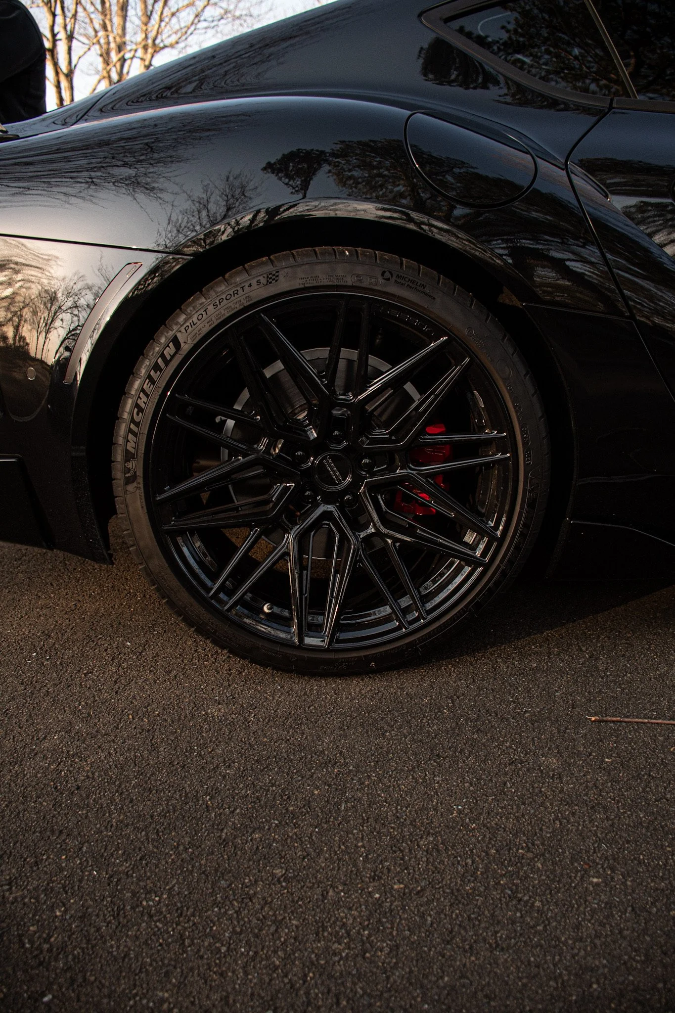 Close-up of a black sports car's front wheel with black alloy rims and a Michelin Pilot Sport 4 S tire, parked on asphalt during sunset with trees reflected on the car's glossy surface.