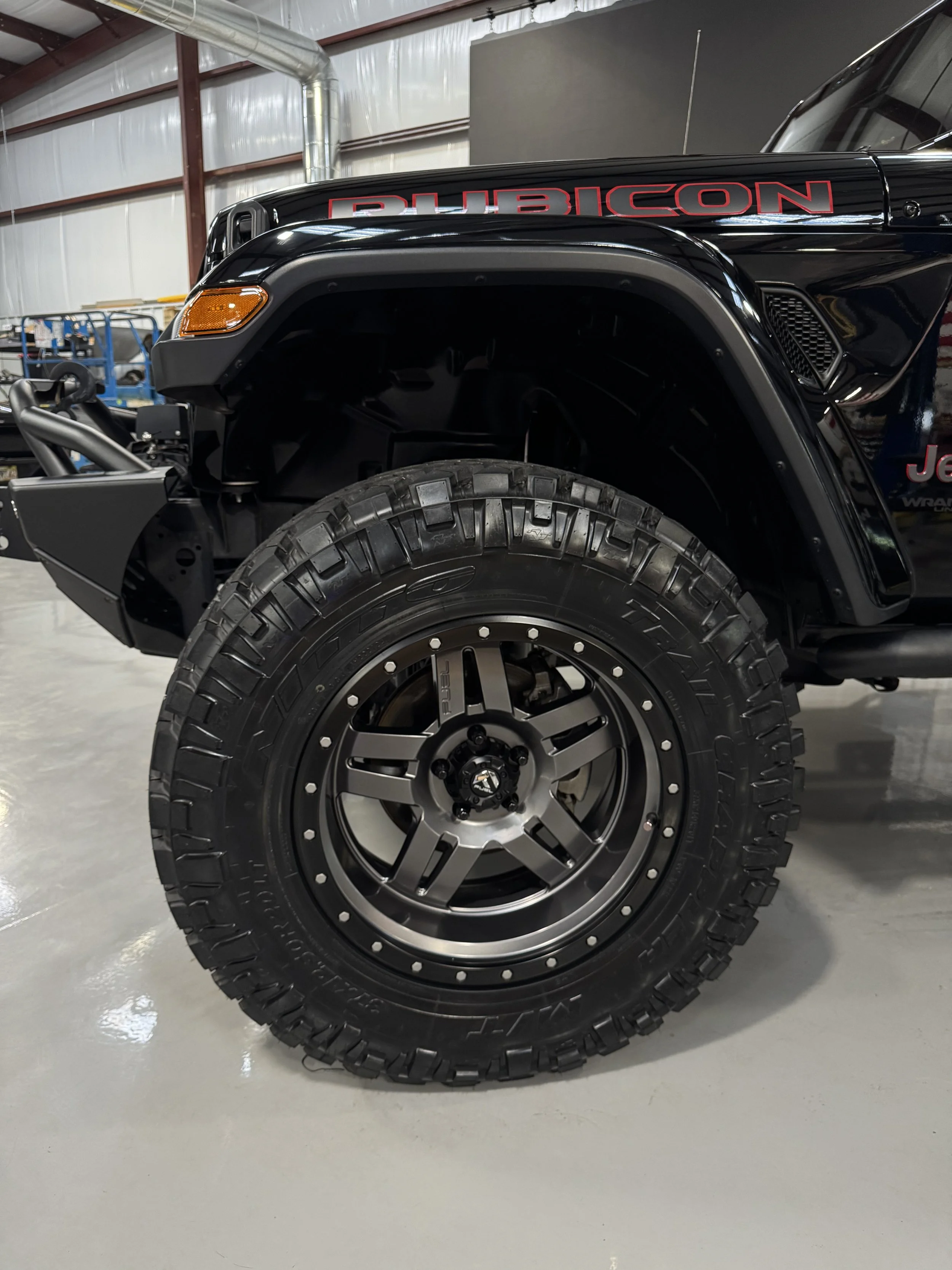 Close-up of a black Jeep Rubicon with large off-road tires and black alloy wheels in a garage or workshop.