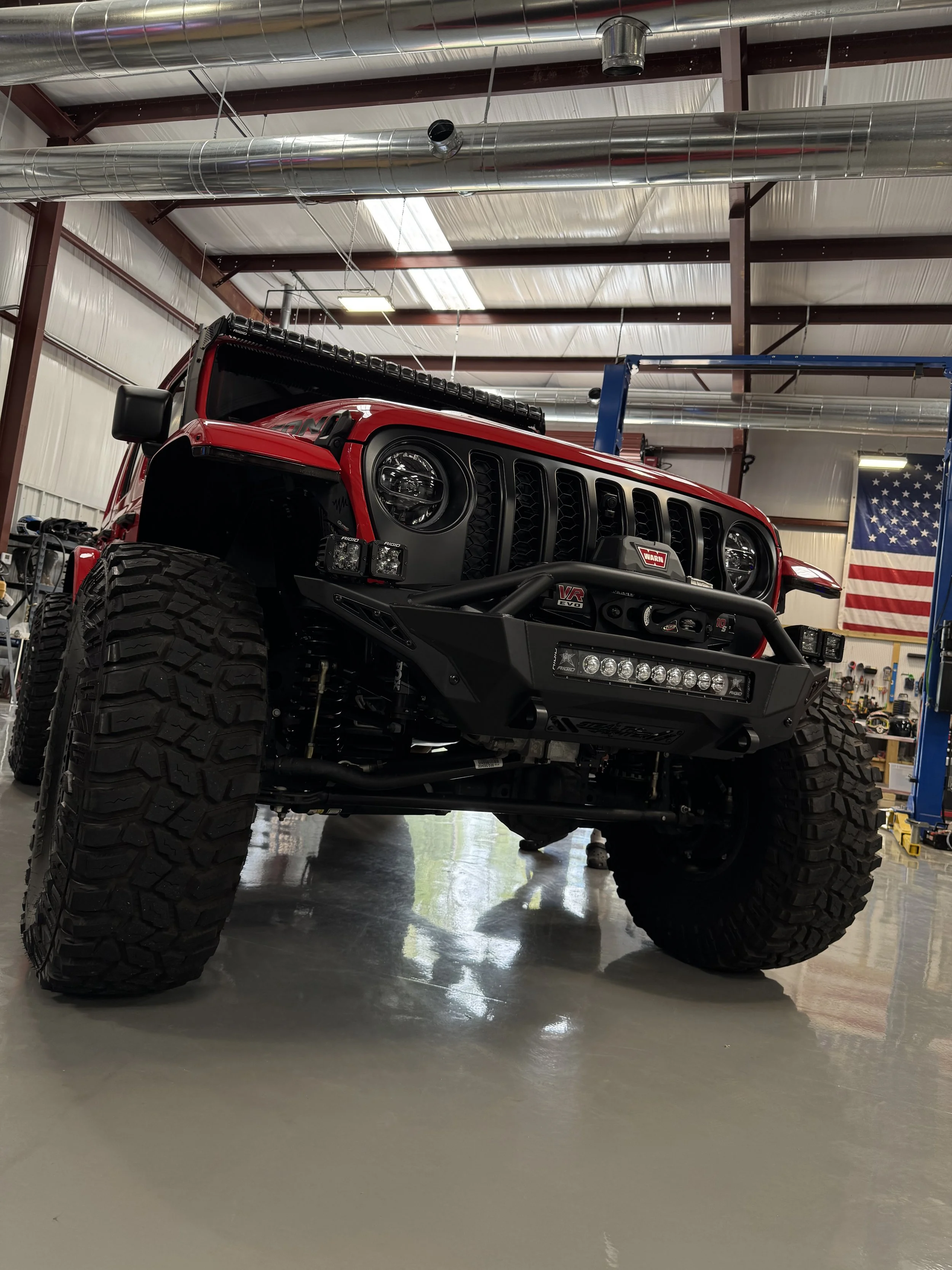 Red off-road vehicle with large tires and black grille inside a garage or workshop, with American flag and industrial pipes overhead.