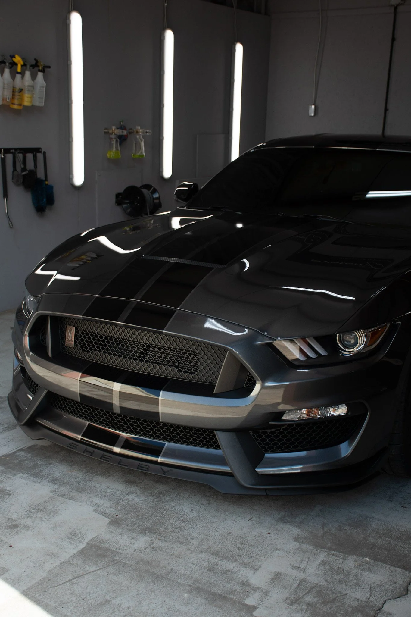 Front of a black sports car with black racing stripes parked indoors, with fluorescent lights and cleaning supplies on the wall behind.