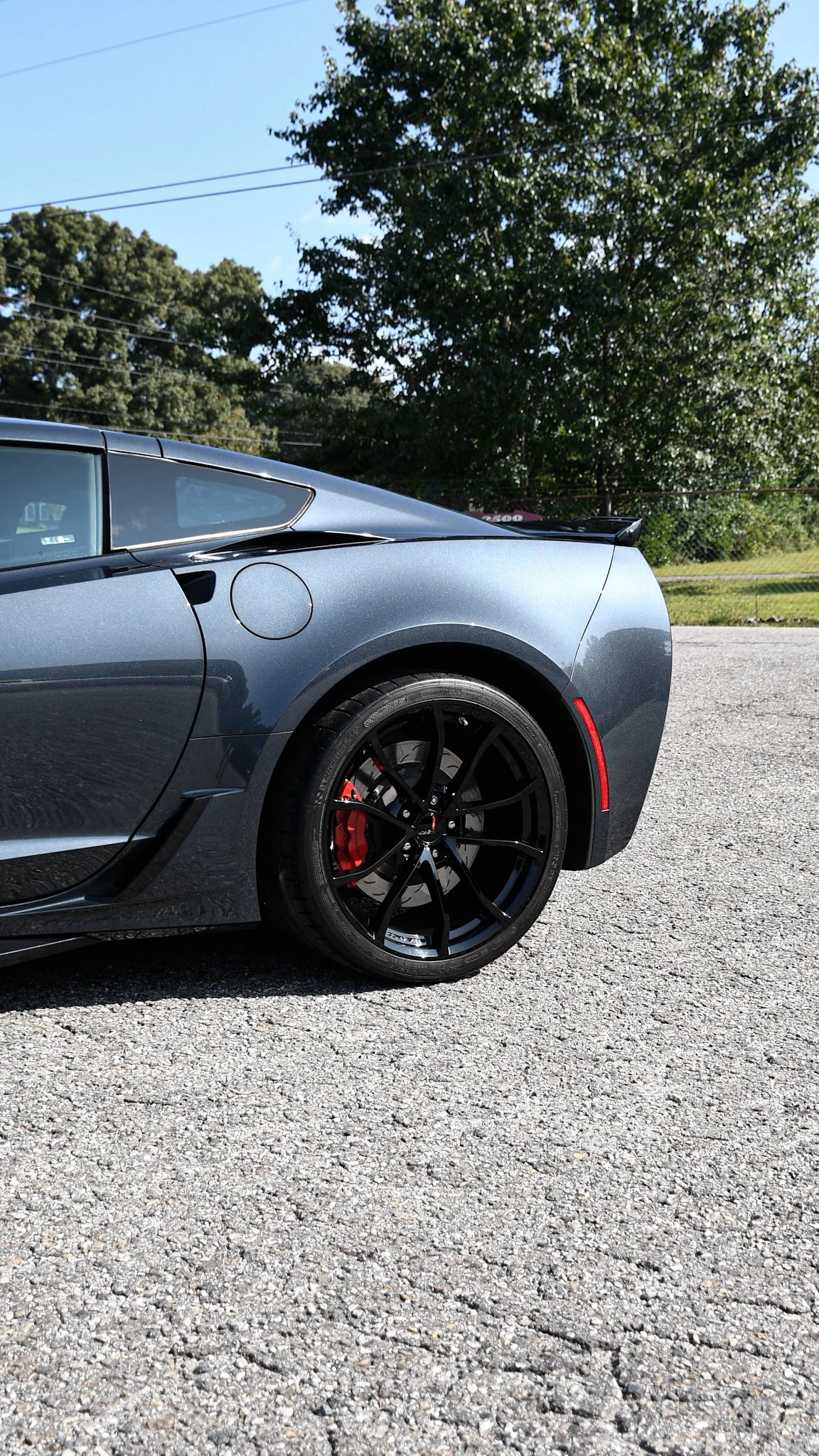 Close-up of the rear side of a gray sports car parked on a gravel surface, with black rims, red brake calipers, a background of trees, and a bright blue sky.