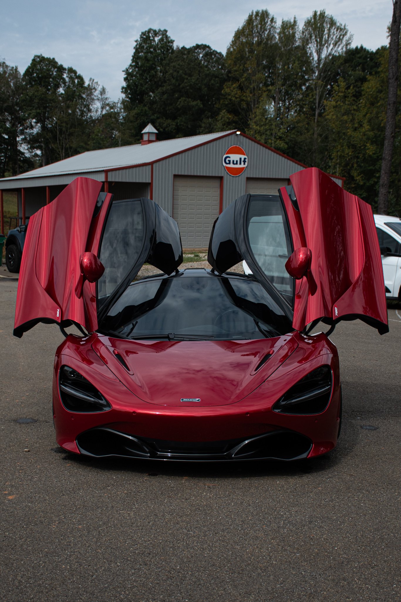 Red sports car with open butterfly doors parked in front of a Gulf gas station building with trees in the background.
