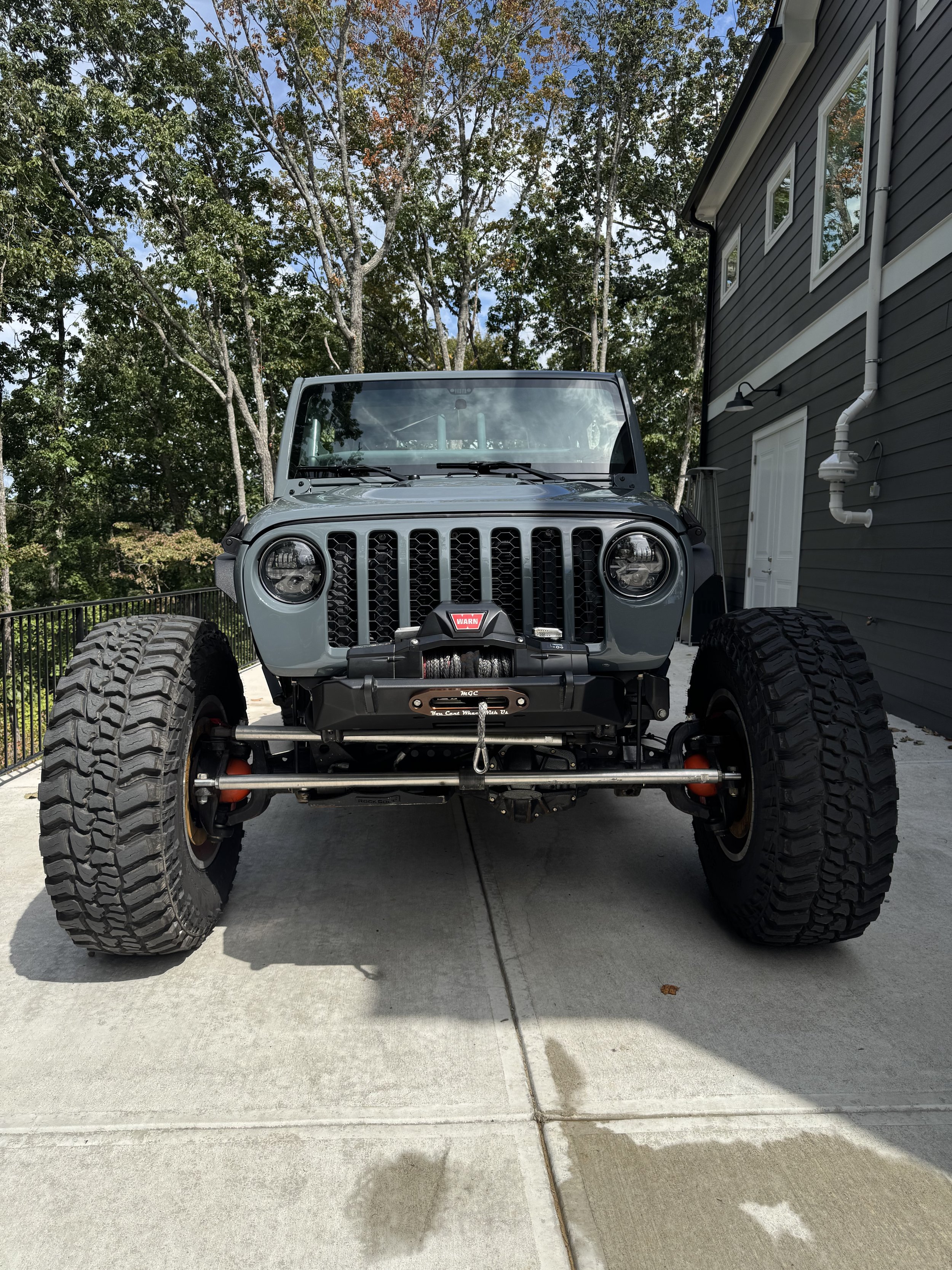 Front view of a gray off-road Jeep with large tires parked on a concrete driveway next to a house with gray siding and white trim, surrounded by trees.