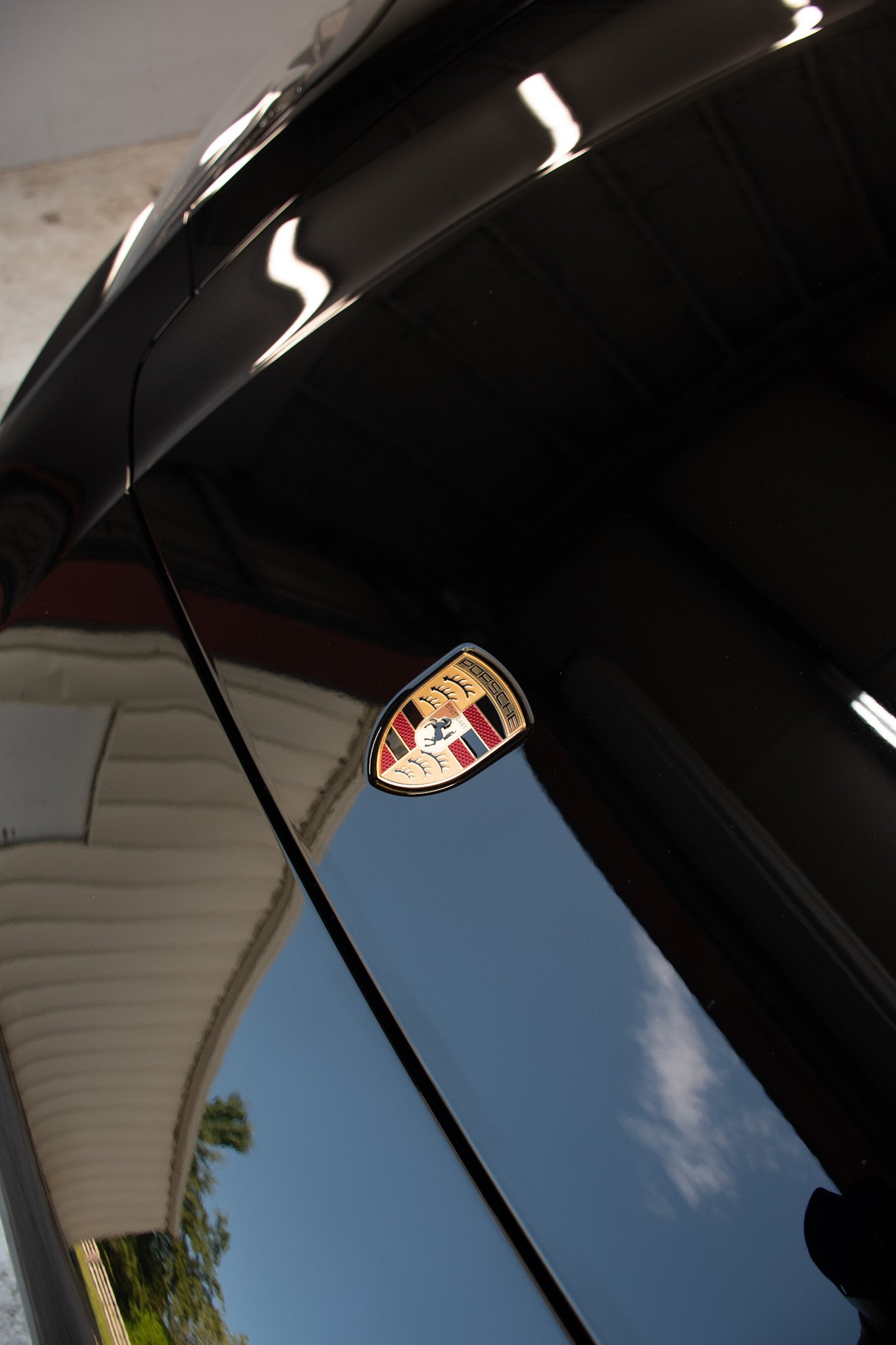 Close-up of a black Porsche car hood with the Porsche emblem and reflections of a white building, blue sky, and trees.