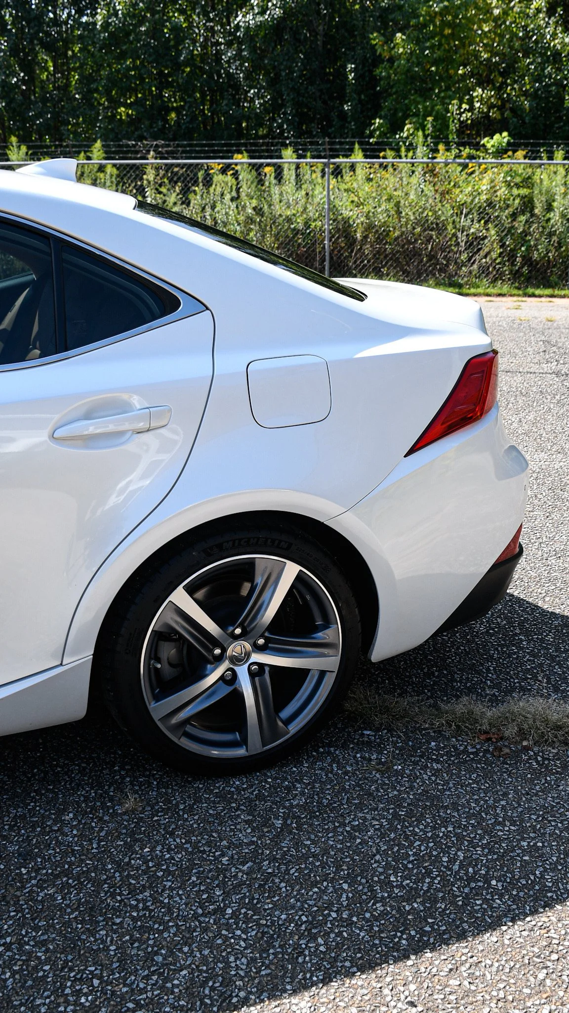 Close-up of the back left side of a white car parked on a gravel surface, with a chain-link fence and green bushes in the background.