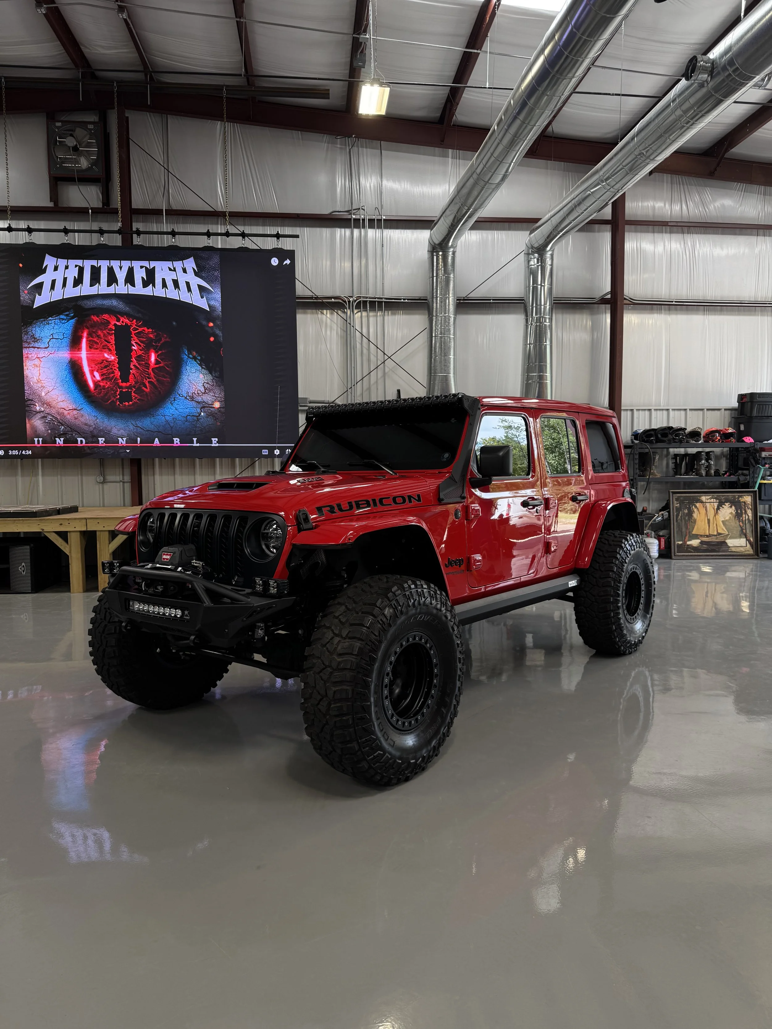 Red Jeep Rubicon on a polished concrete floor inside a large garage or warehouse with a metal roof and exposed ductwork. A large screen displays art with the word 'Helldivers' and a large eye with a red iris.