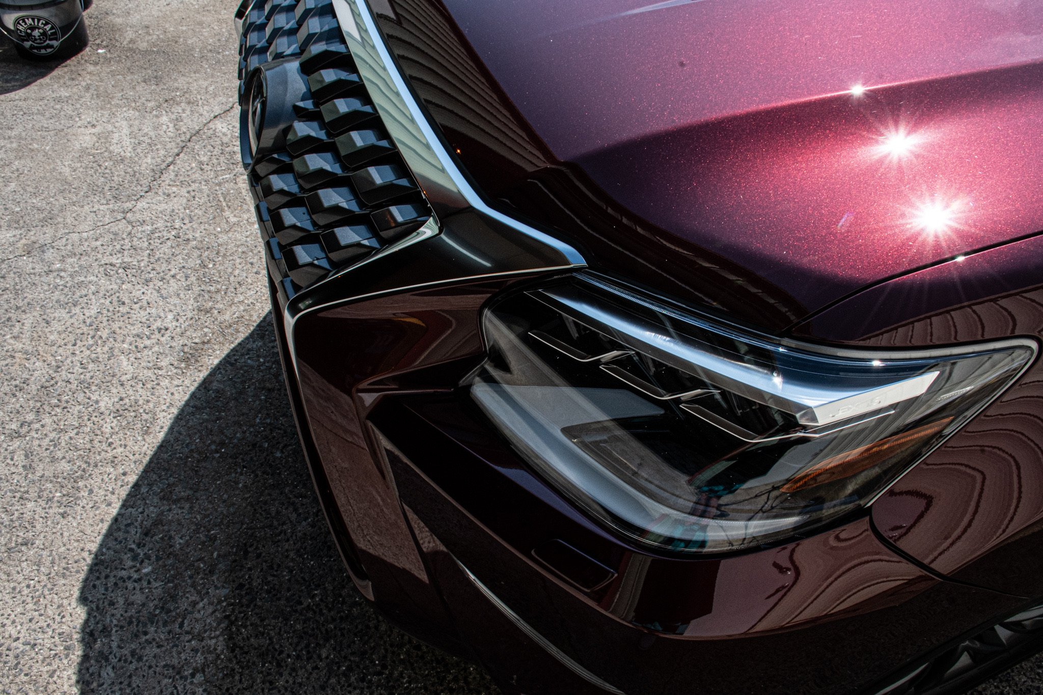 Close-up of the front right side of a shiny, dark-colored car with a metallic finish, showing the headlight, grille, and part of the hood, parked on a textured concrete surface.