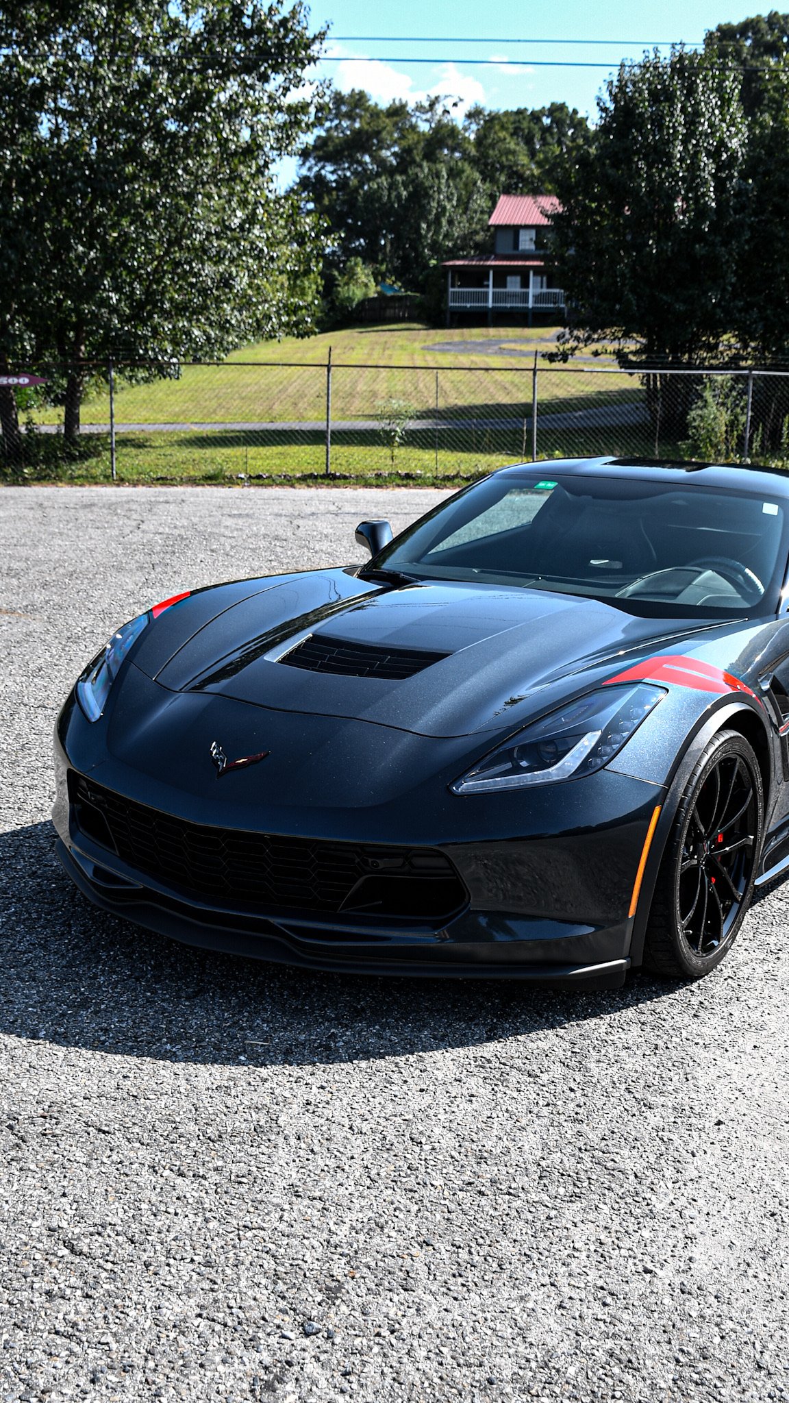 Black Chevrolet Corvette sports car with red accents parked on gravel with a fenced grassy area, trees, and a house with a porch in the background.