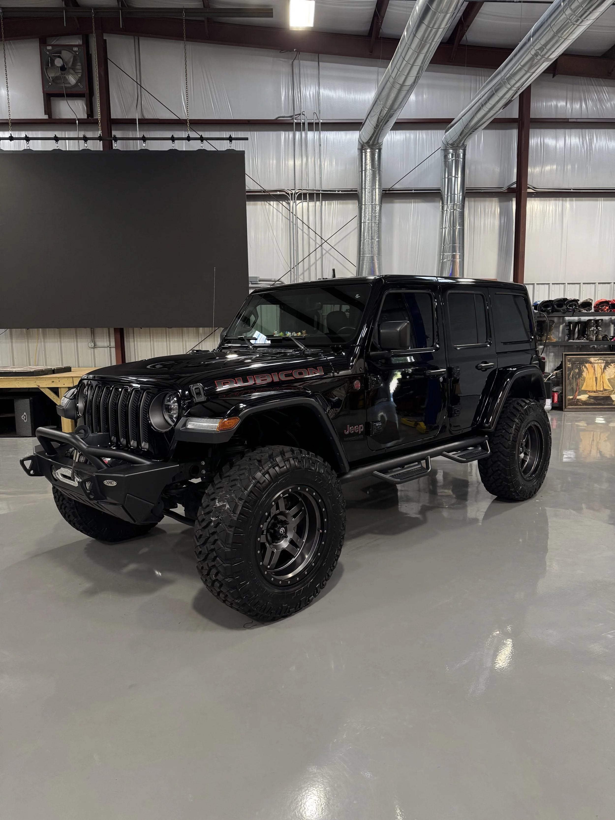 A black Jeep Rubicon parked inside a large industrial-style garage or workshop. The vehicle has large off-road tires, black rims, and is slightly lifted. In the background, there are metal ductwork, a large gray display or screen, shelves with helmet