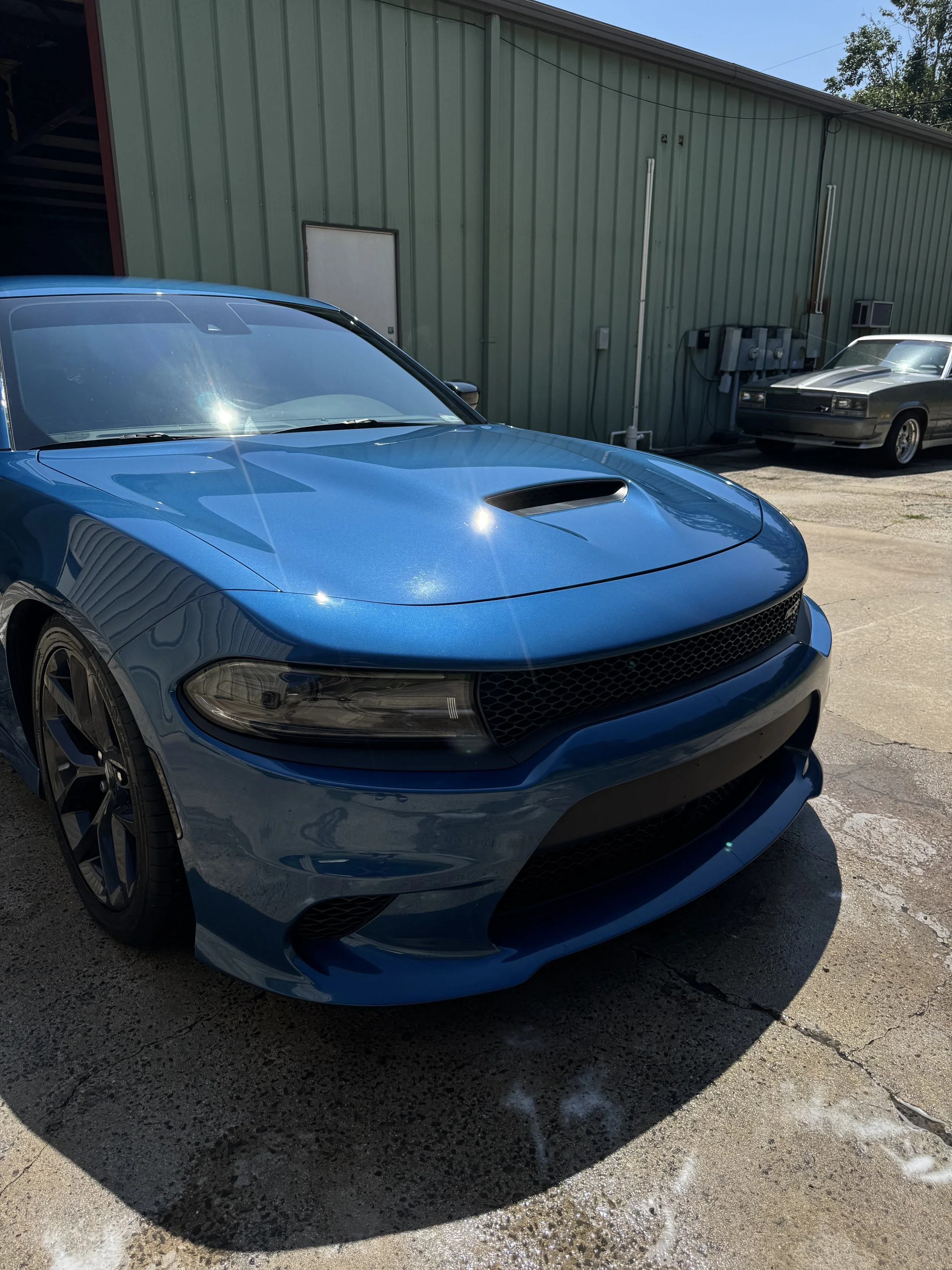 A blue sports car parked in front of a green industrial building with another car visible in the background.