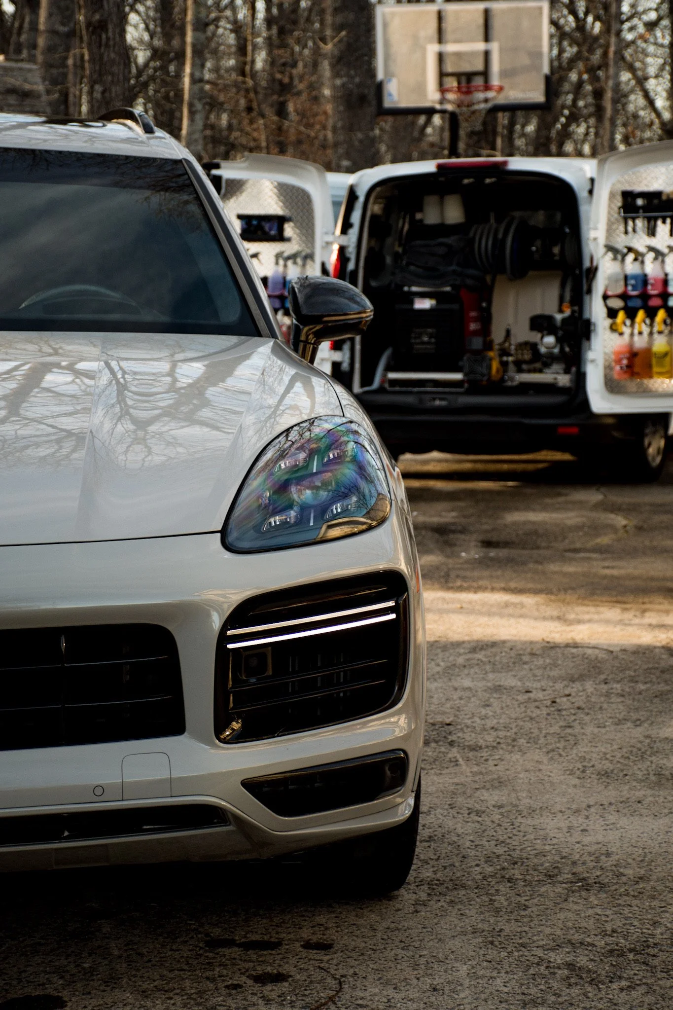 Close-up of front part of a white luxury car parked outdoors, with a toolbox truck and basketball hoop in the background.