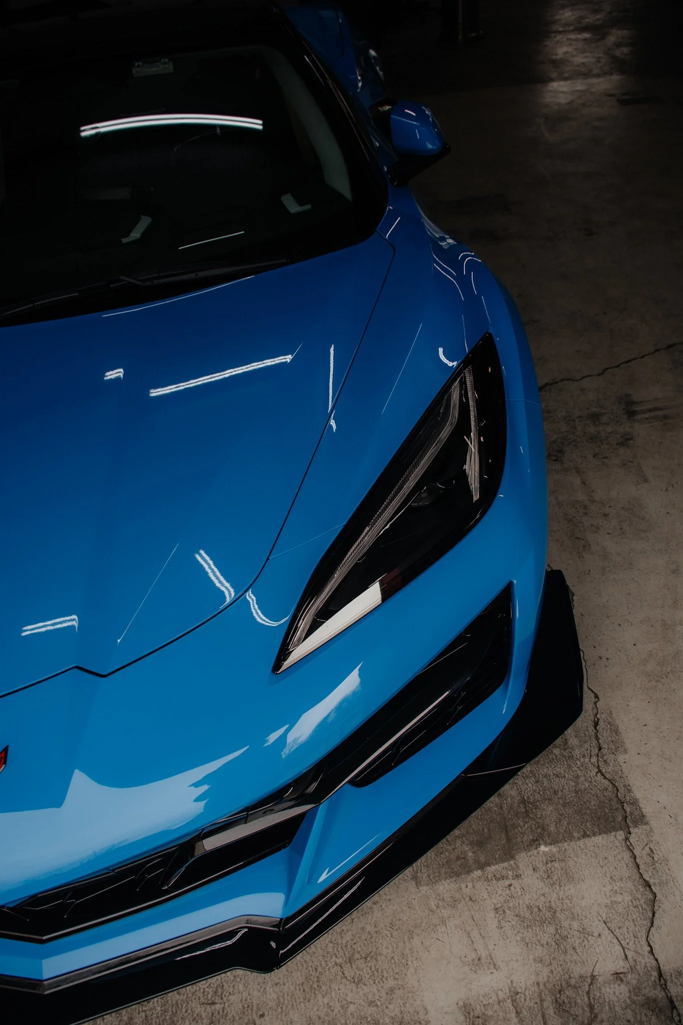 Close-up of the front part of a blue sports car with a sleek design, sharp lines, and an angular headlight, parked on a concrete floor.