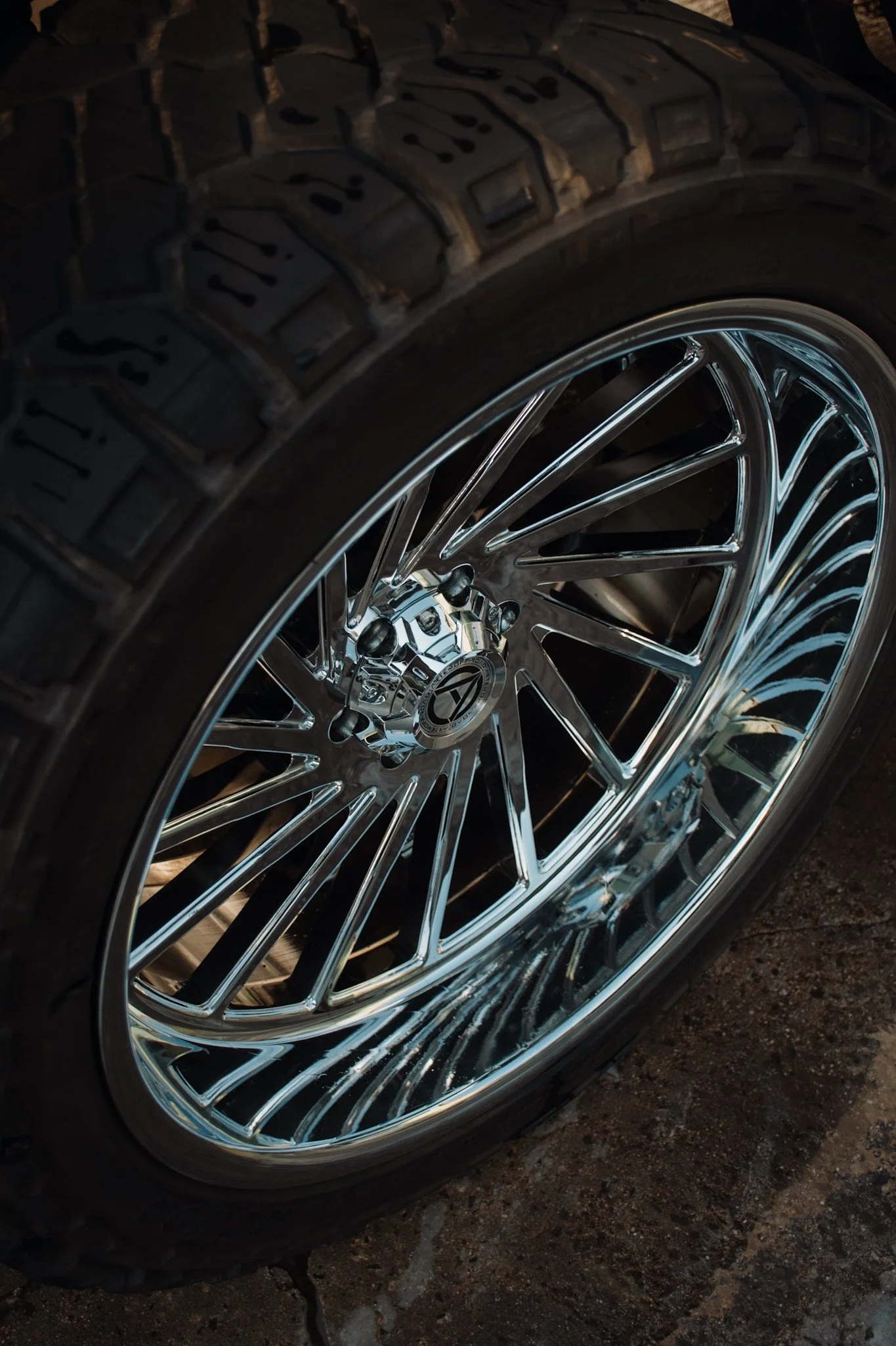A close-up of a car wheel with a shiny chrome rim and a thick, textured tire, resting on a dirt surface.