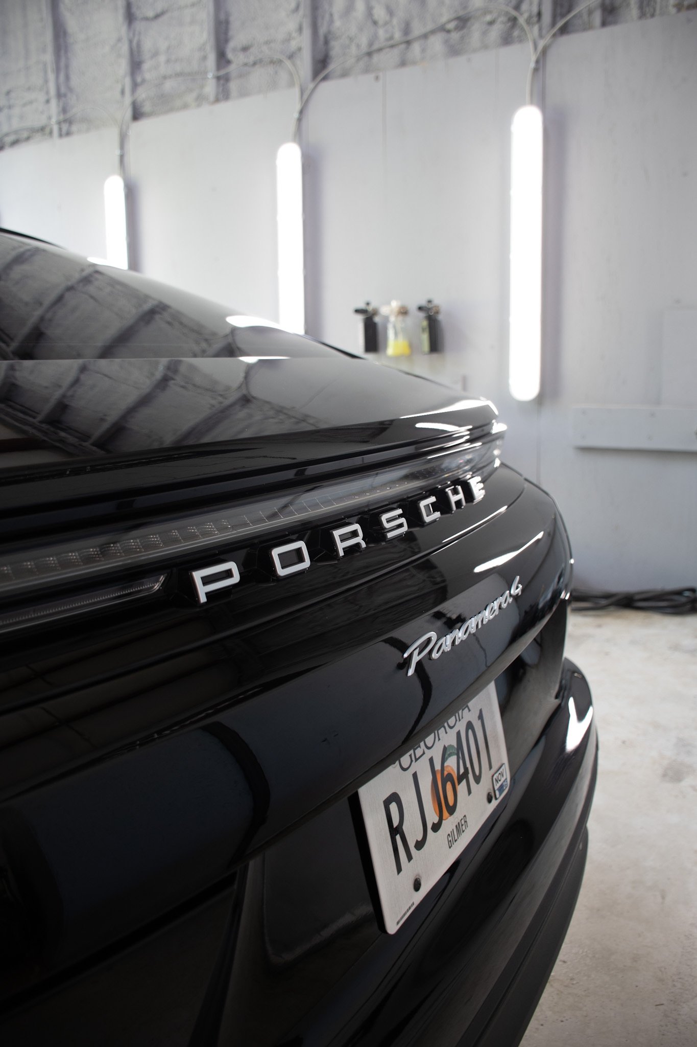 The rear of a black Porsche Panamera with a California license plate is parked inside a garage or showroom. The Porsche logo is visible on the back of the car, and the license plate reads 'RJ6U40.' The background includes white walls and vertical flu